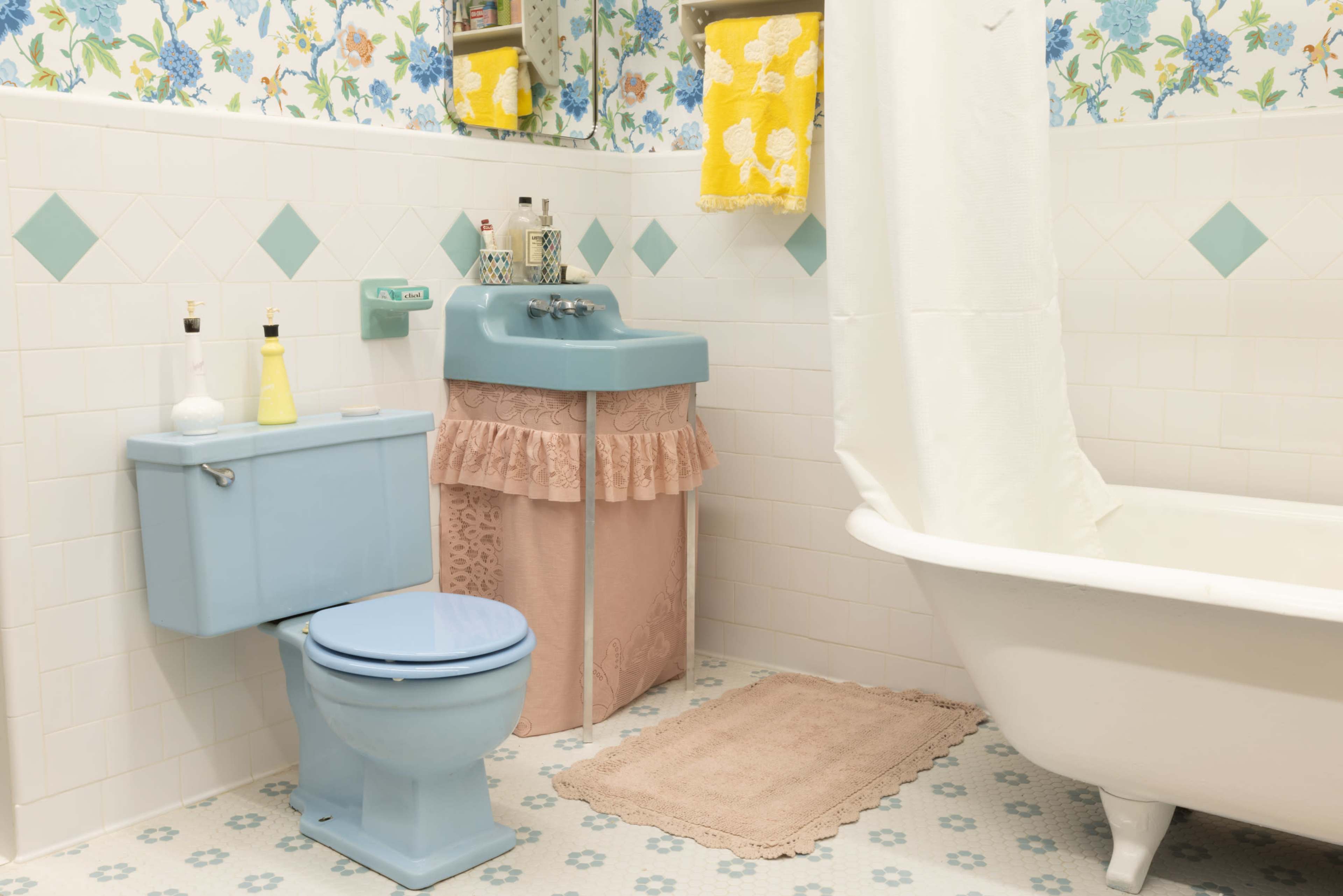 A vintage-themed bathroom featuring a pale blue toilet and sink, a white clawfoot bathtub, and floral wallpaper with decorative elements.