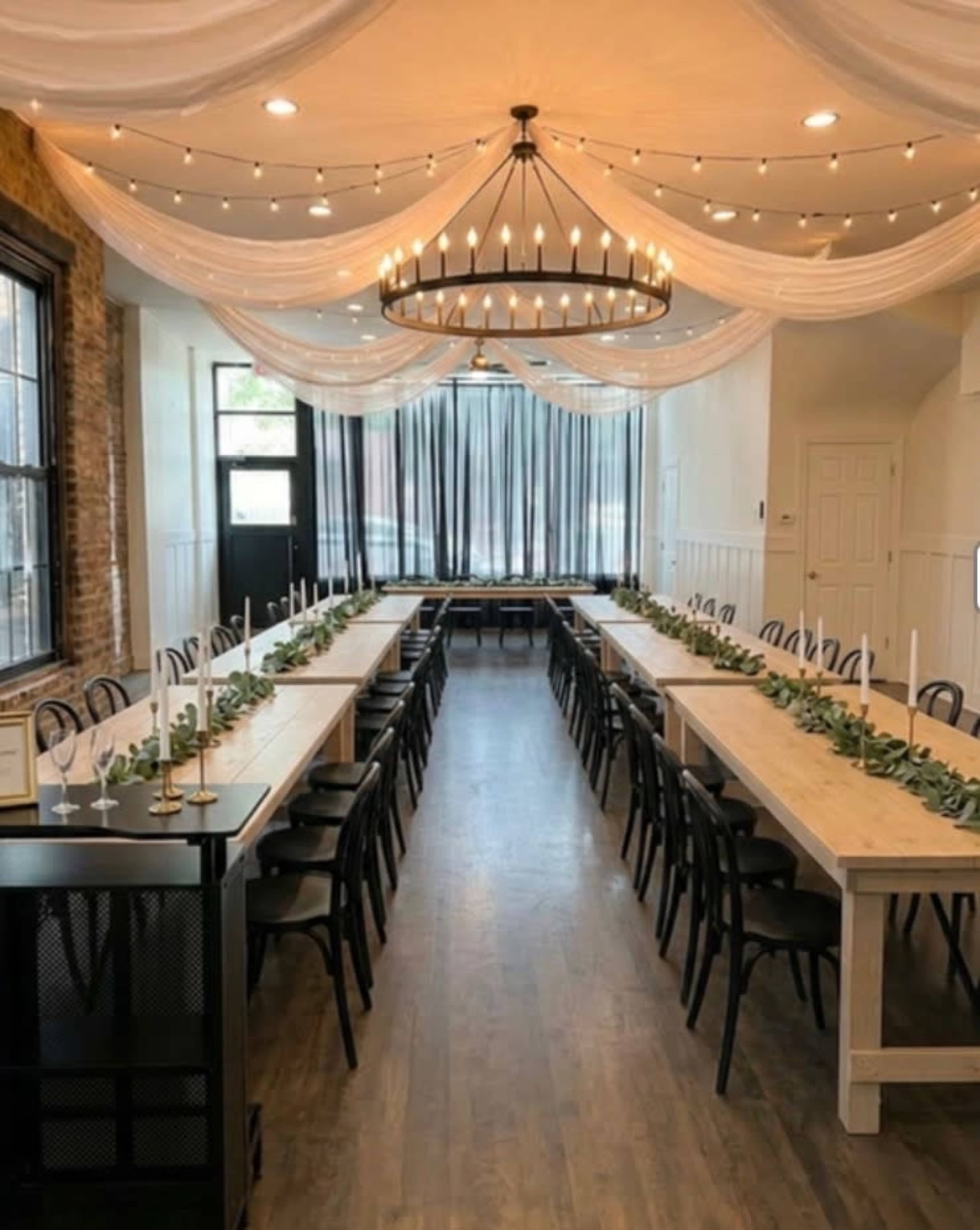 The image shows a banquet hall setup with long tables arranged in two parallel rows, adorned with greenery and candleholders, beneath a chandelier and draped fabric on the ceiling.