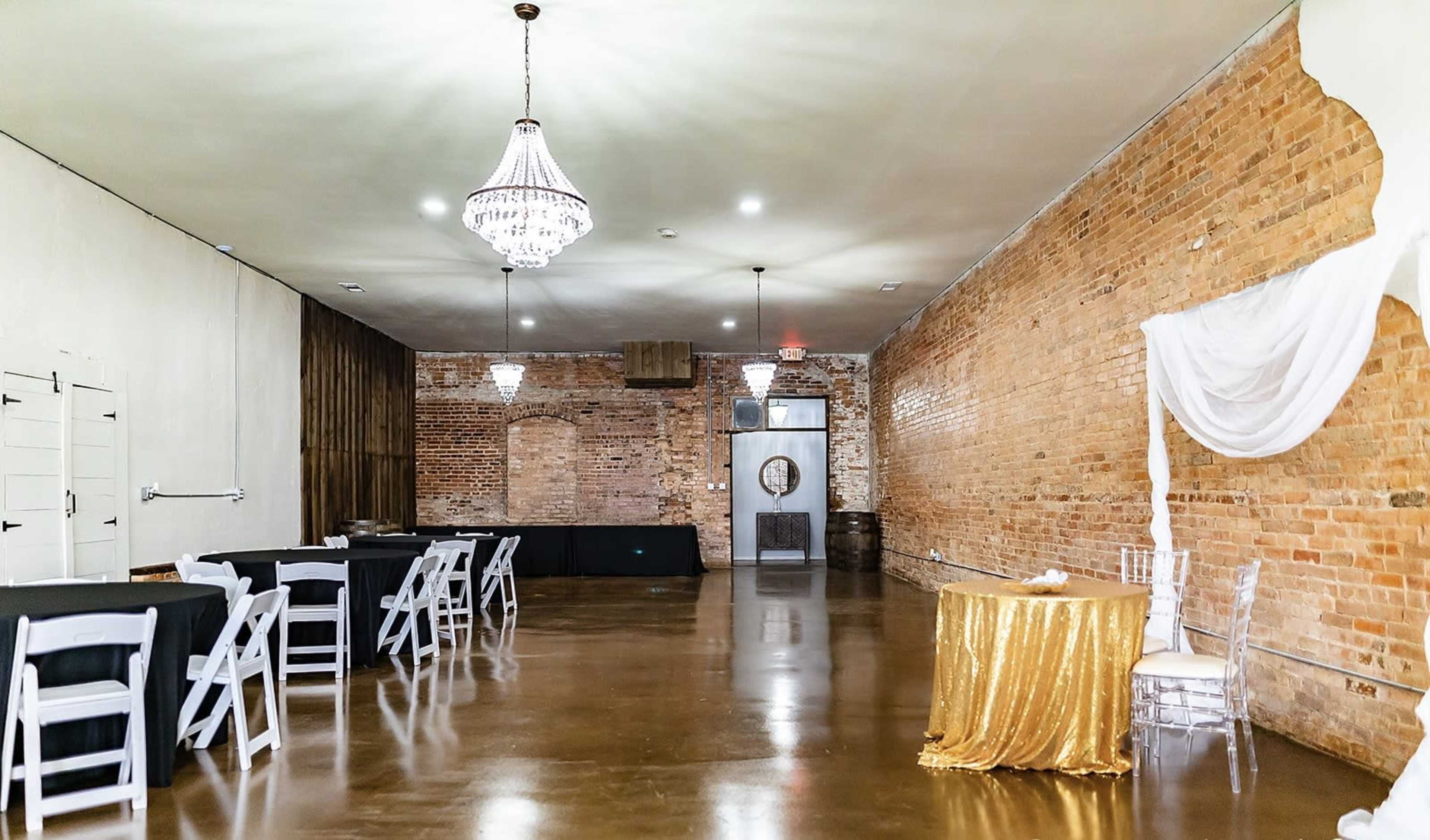 The image shows a spacious, well-lit event room featuring brick walls, a polished concrete floor, black tables, and a decorative gold tablecloth.