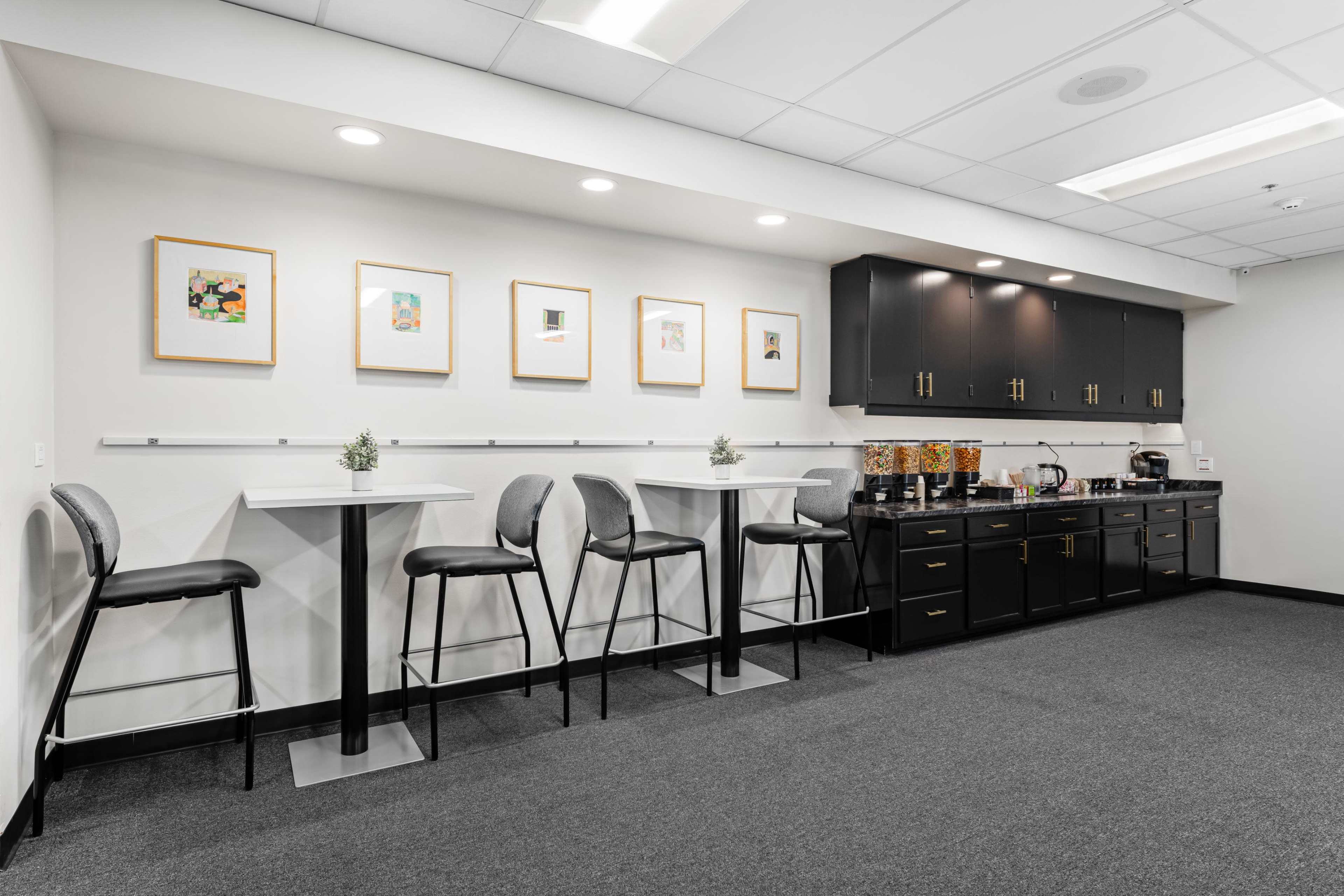 The image depicts a modern break room with a series of black cabinets, a coffee station, and high tables with stools arranged along the wall.