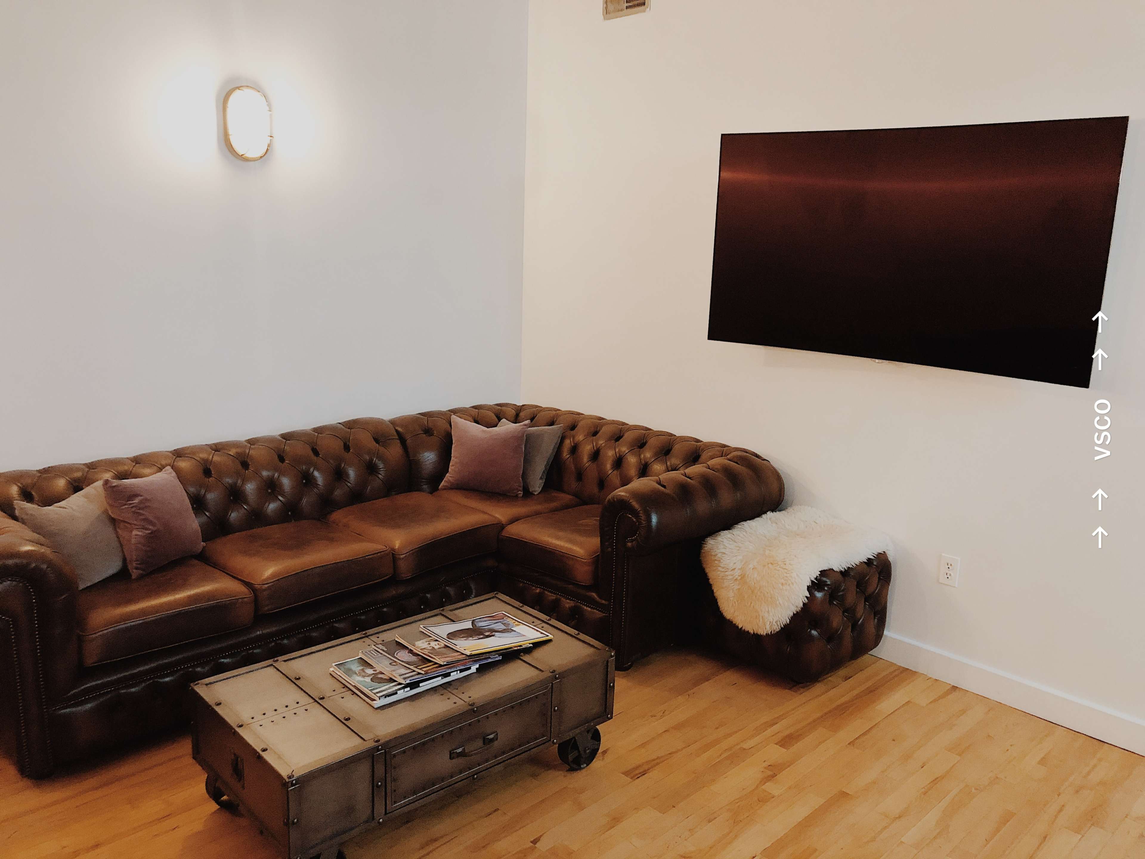 A living room featuring a brown leather Chesterfield sofa with cushions, a rectangular wooden coffee table, and a wall-mounted flat-screen TV.