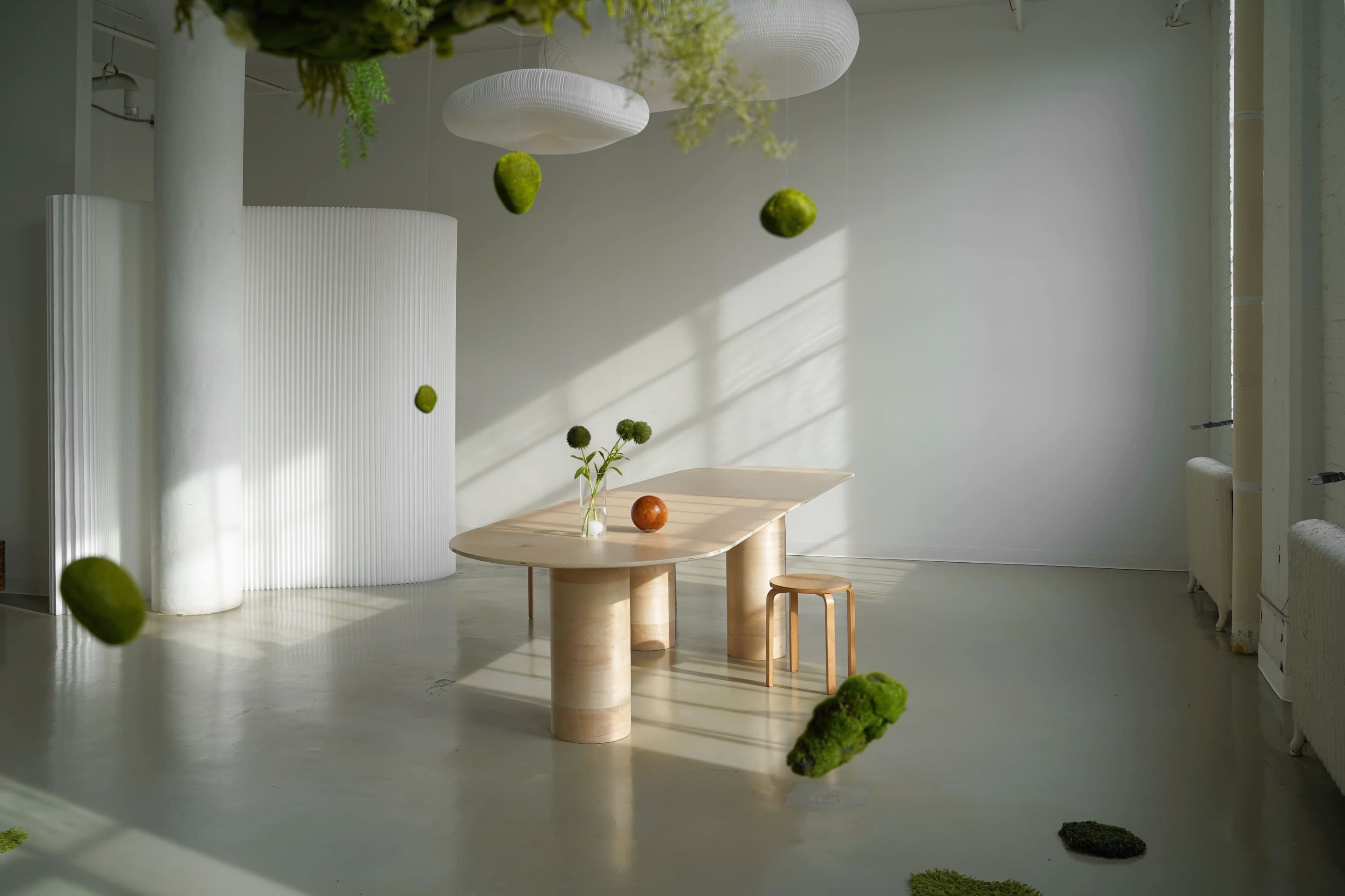A light-filled room features a minimalist wooden table, a chair, and suspended green moss spheres, creating an open and airy atmosphere.