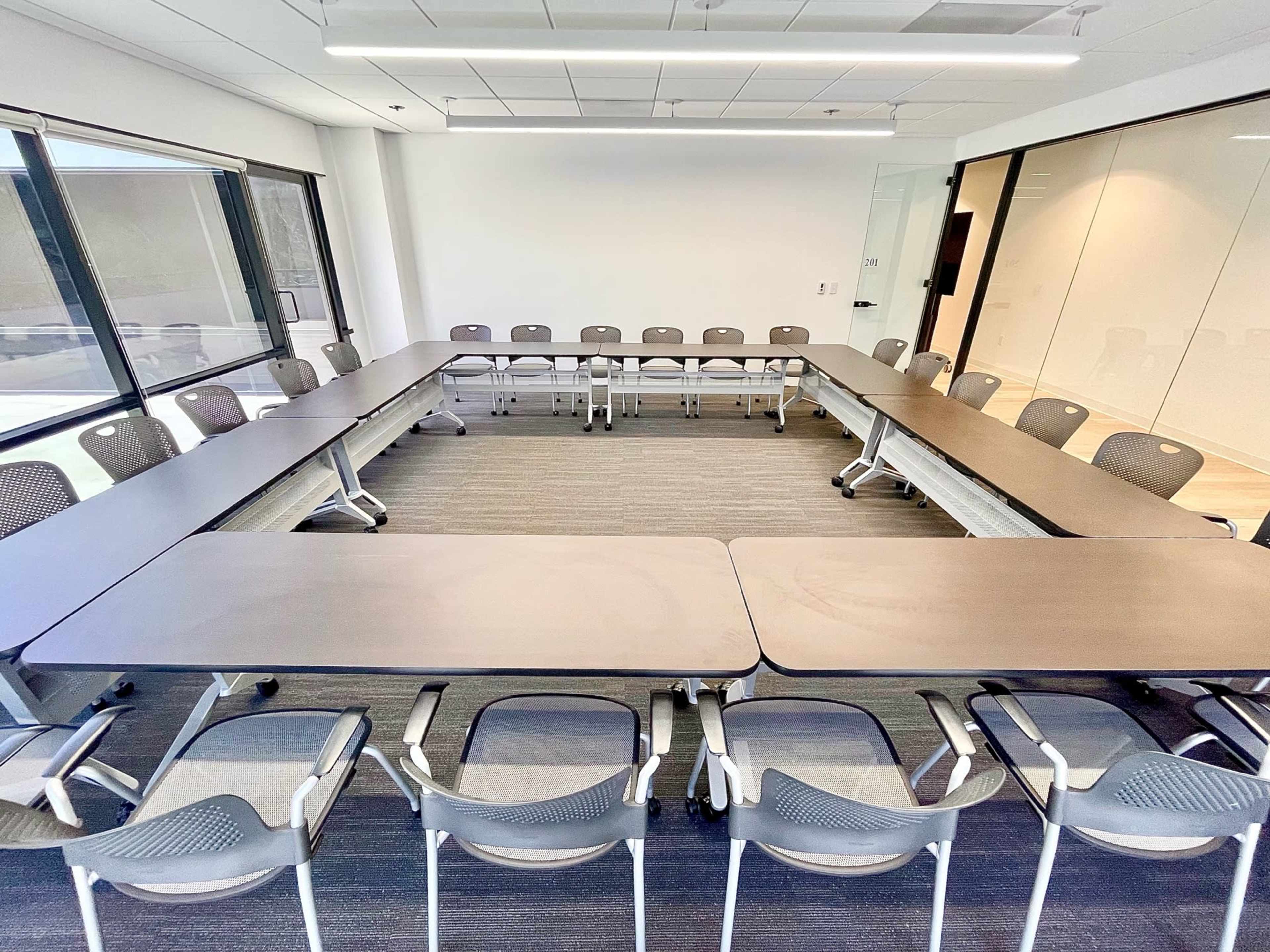 A conference room features a large rectangular arrangement of tables surrounded by chairs, with large windows along one wall.