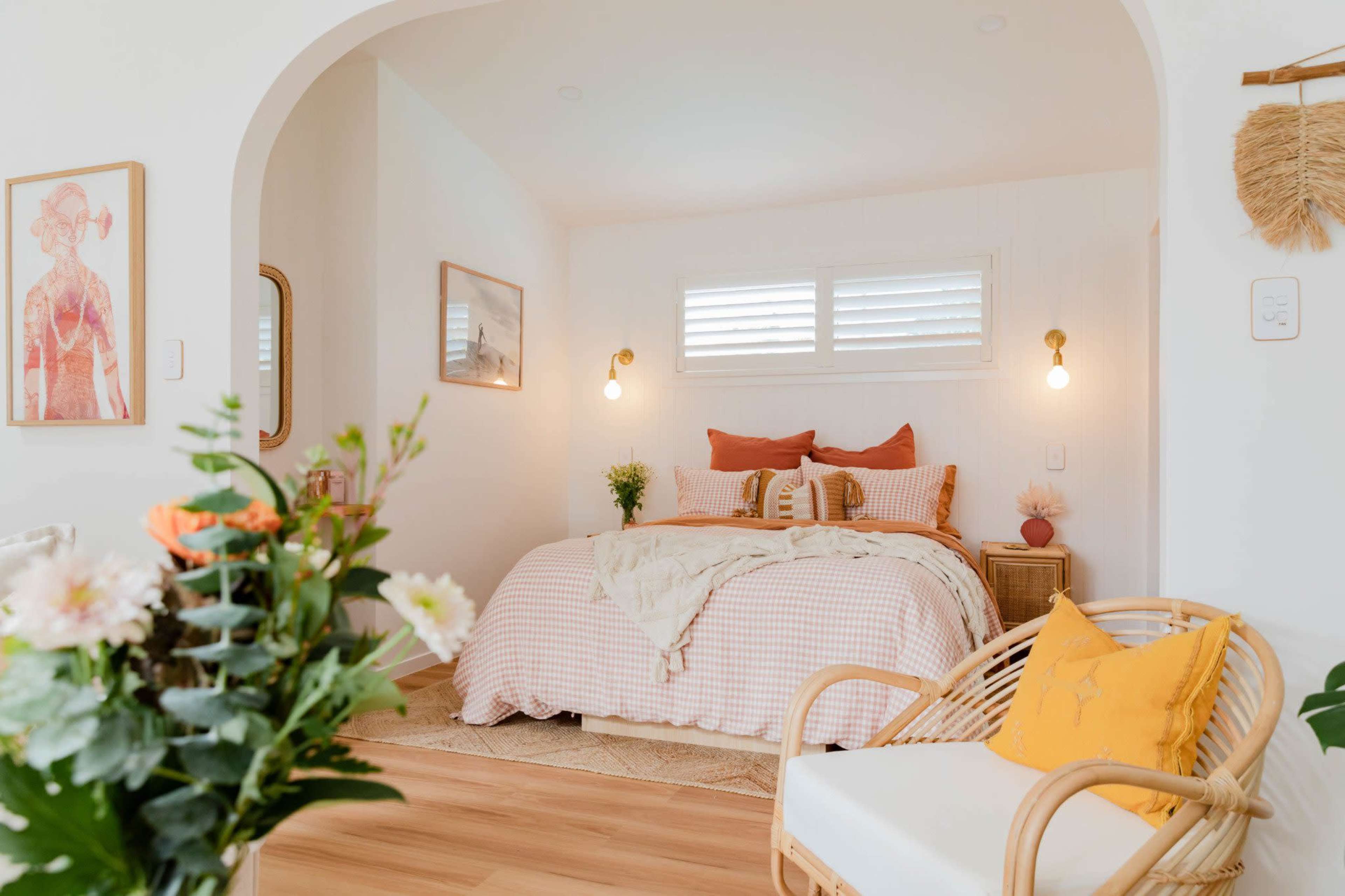 The image shows a cozy bedroom with a checkered bedspread, decorative pillows, and natural light filtering through the windows beside a small seating area and a floral arrangement in the foreground.