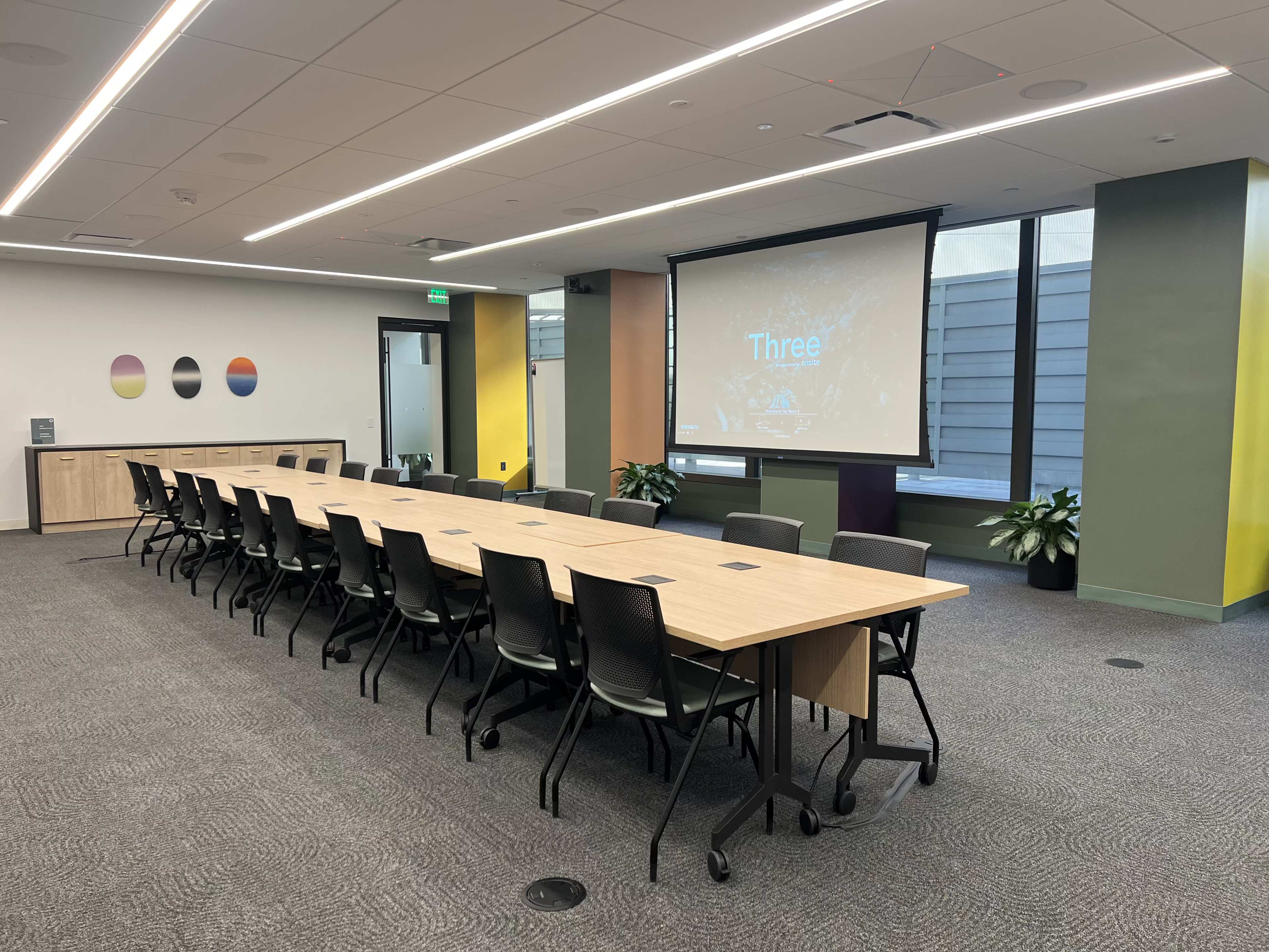 A large conference room is set up with a long table and multiple chairs, featuring a projector screen displaying the word "Three."