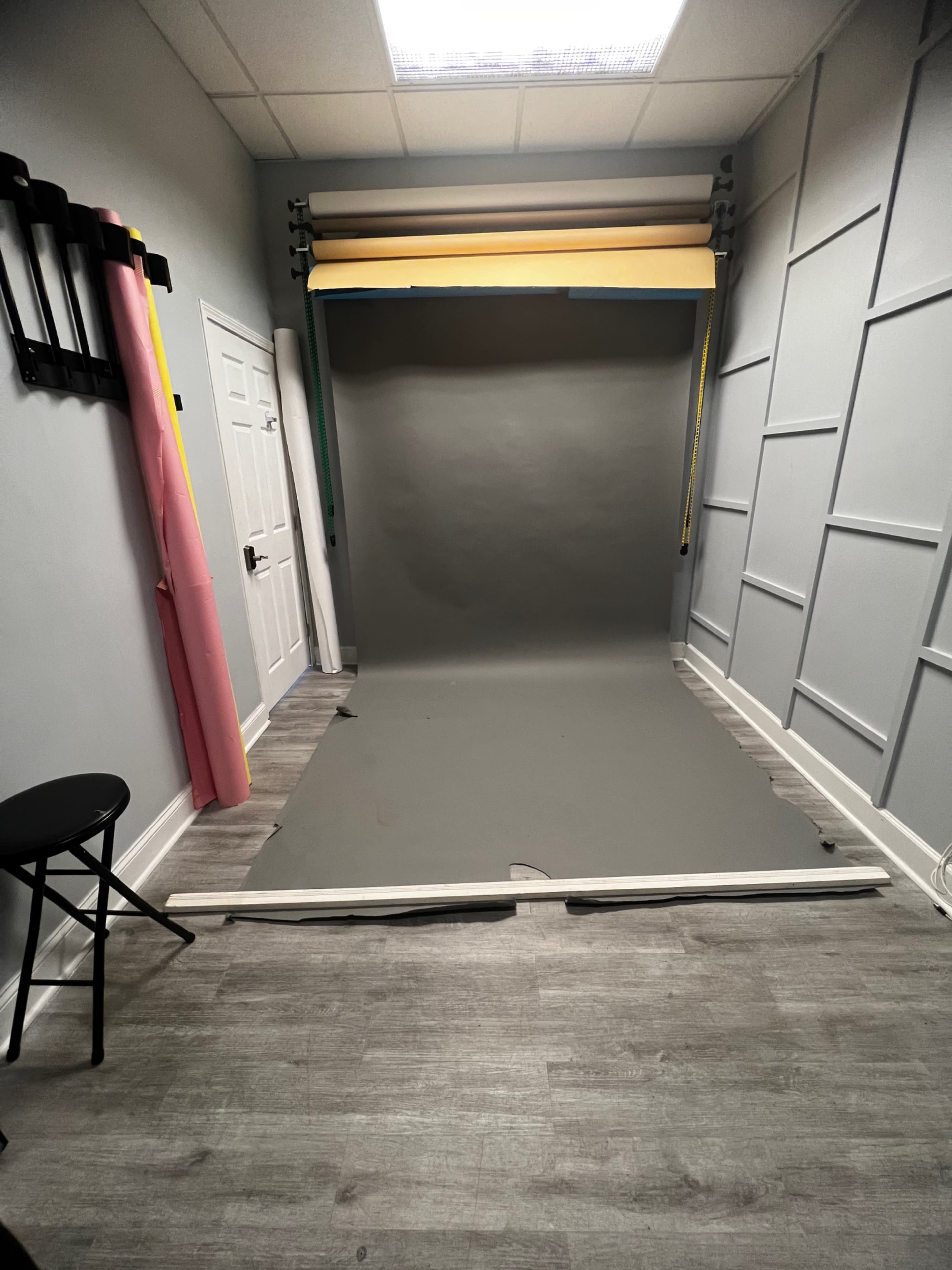 A small photography studio with a gray backdrop, a black stool, and several rolls of colored paper against the wall.