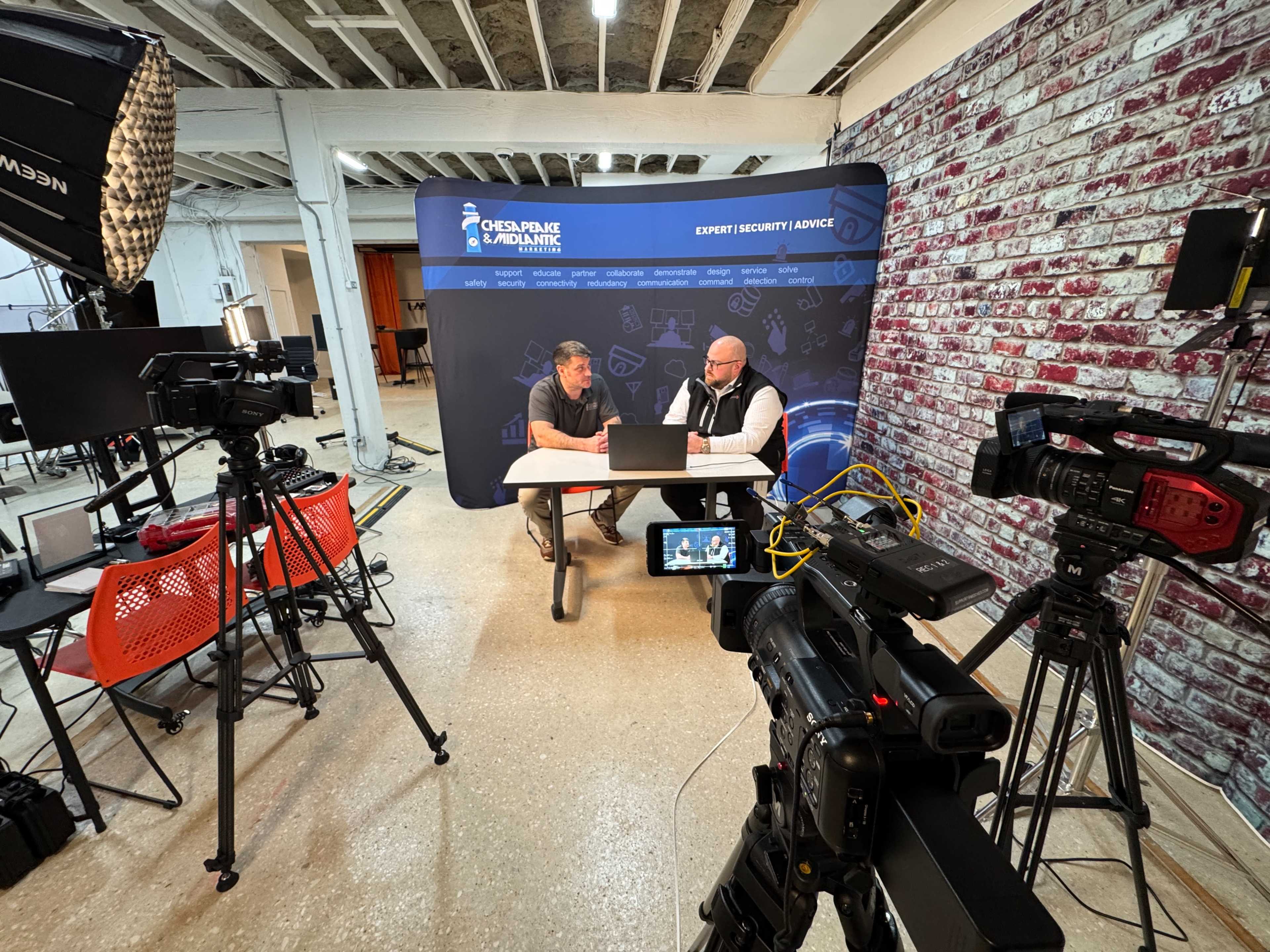 Two men are sitting at a table, engaged in a discussion, with multiple cameras and lighting equipment set up around them in a studio environment.