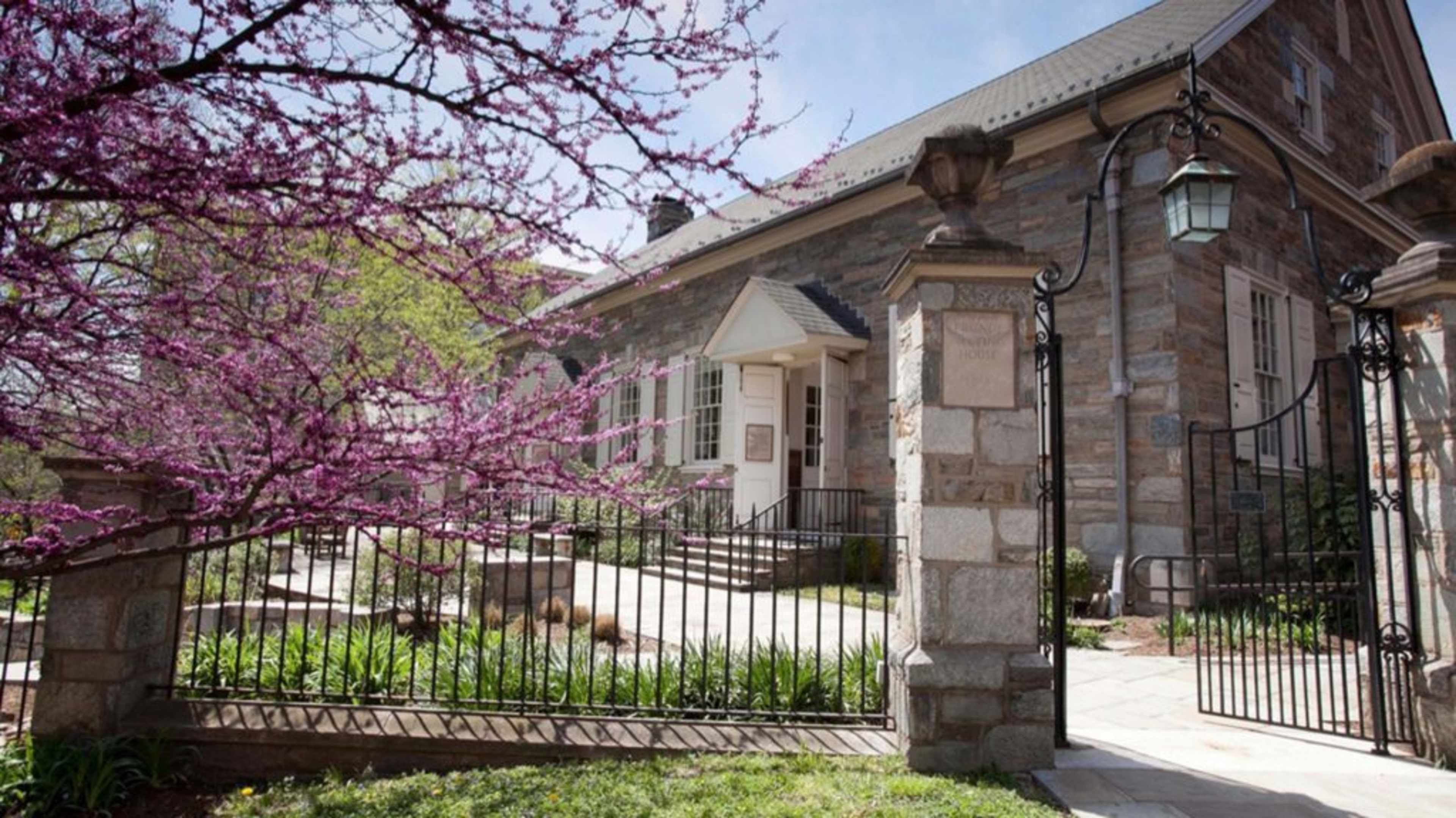 The image shows a stone building with a white entrance, surrounded by blooming pink trees and a wrought iron gate.
