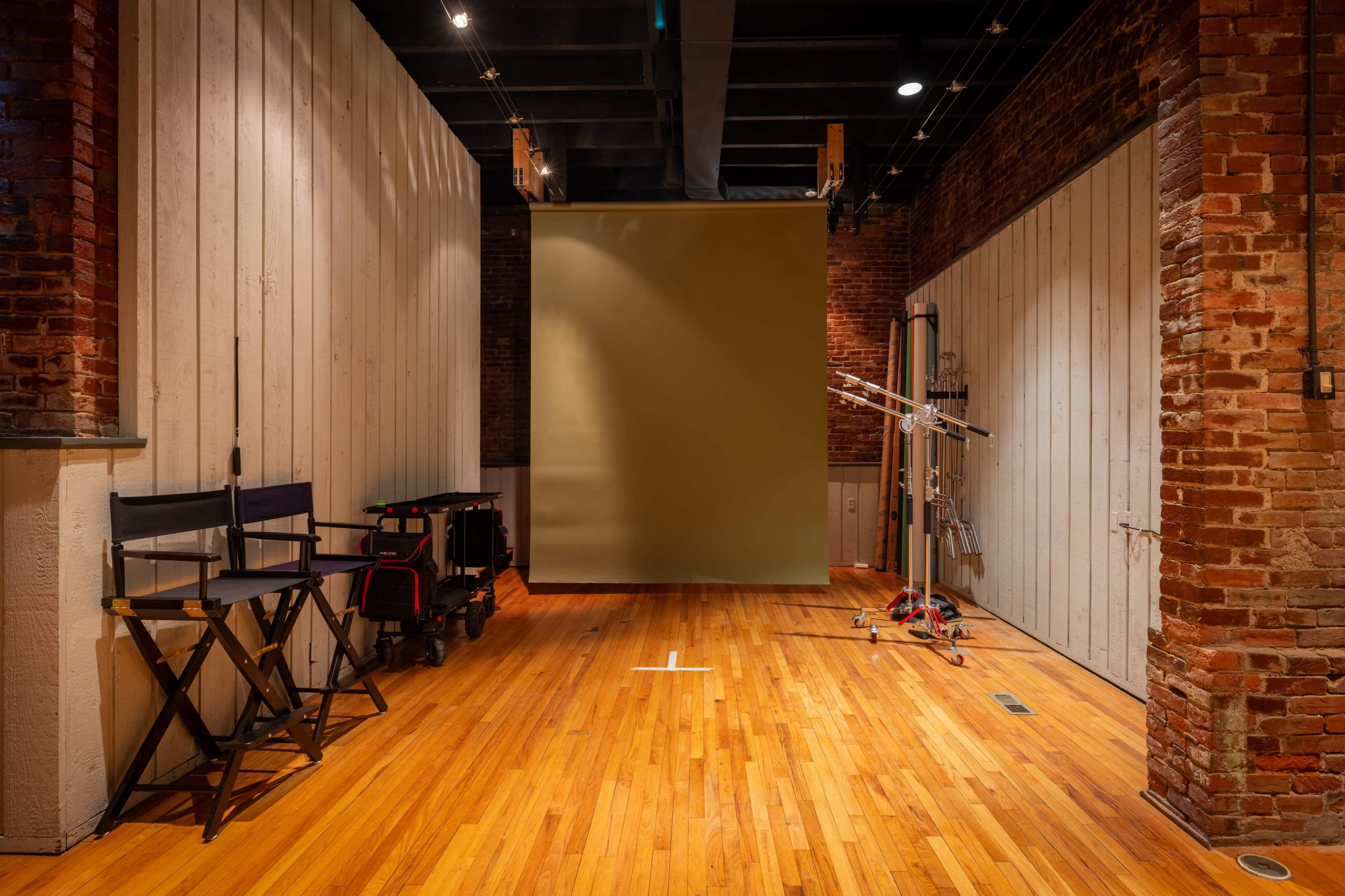 The image shows a spacious photography studio with wooden floors, exposed brick walls, a large backdrop, and various equipment set against one side.