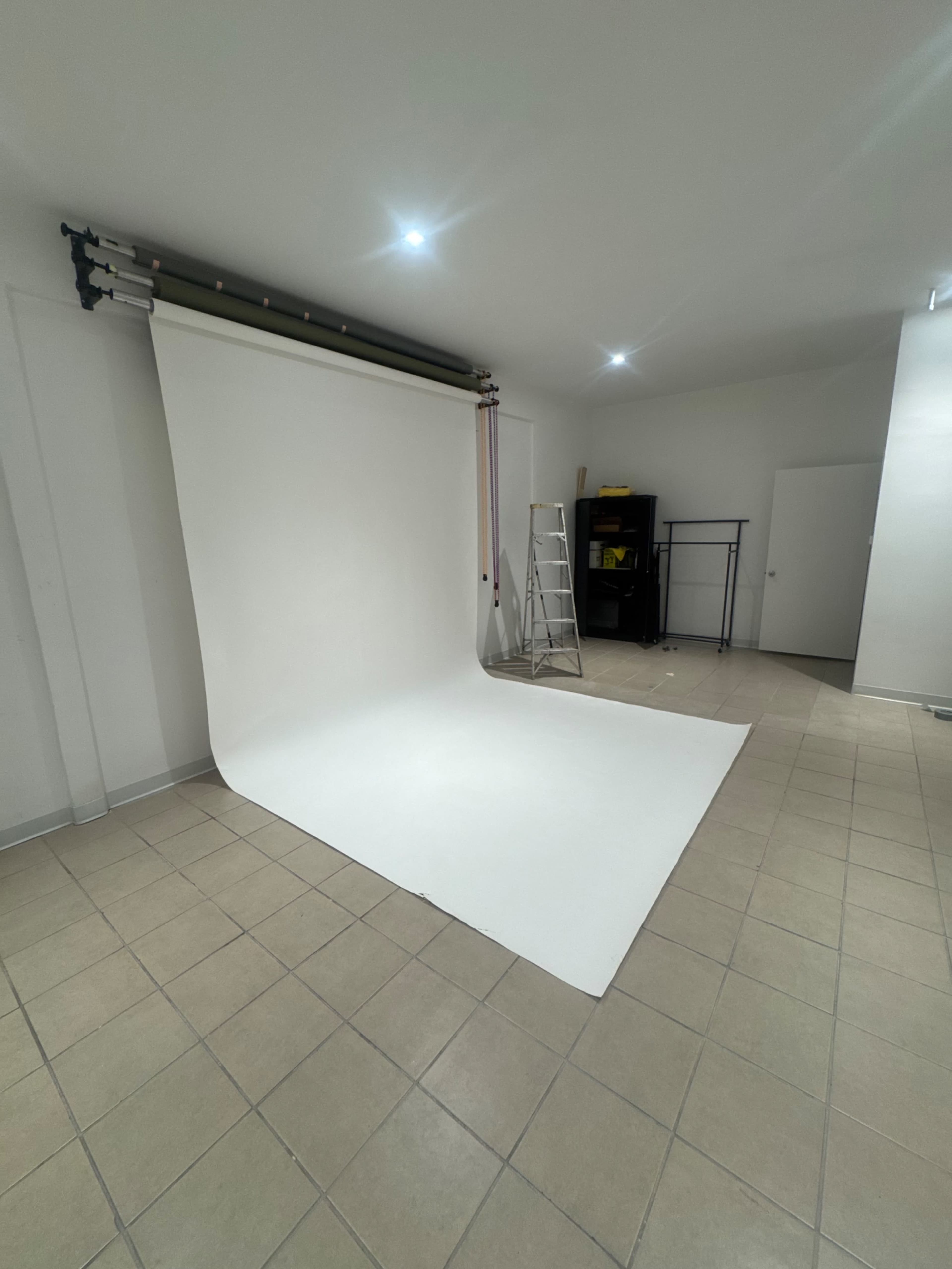 The image shows a photography studio with a white backdrop, a ladder, and a black storage rack against the wall.