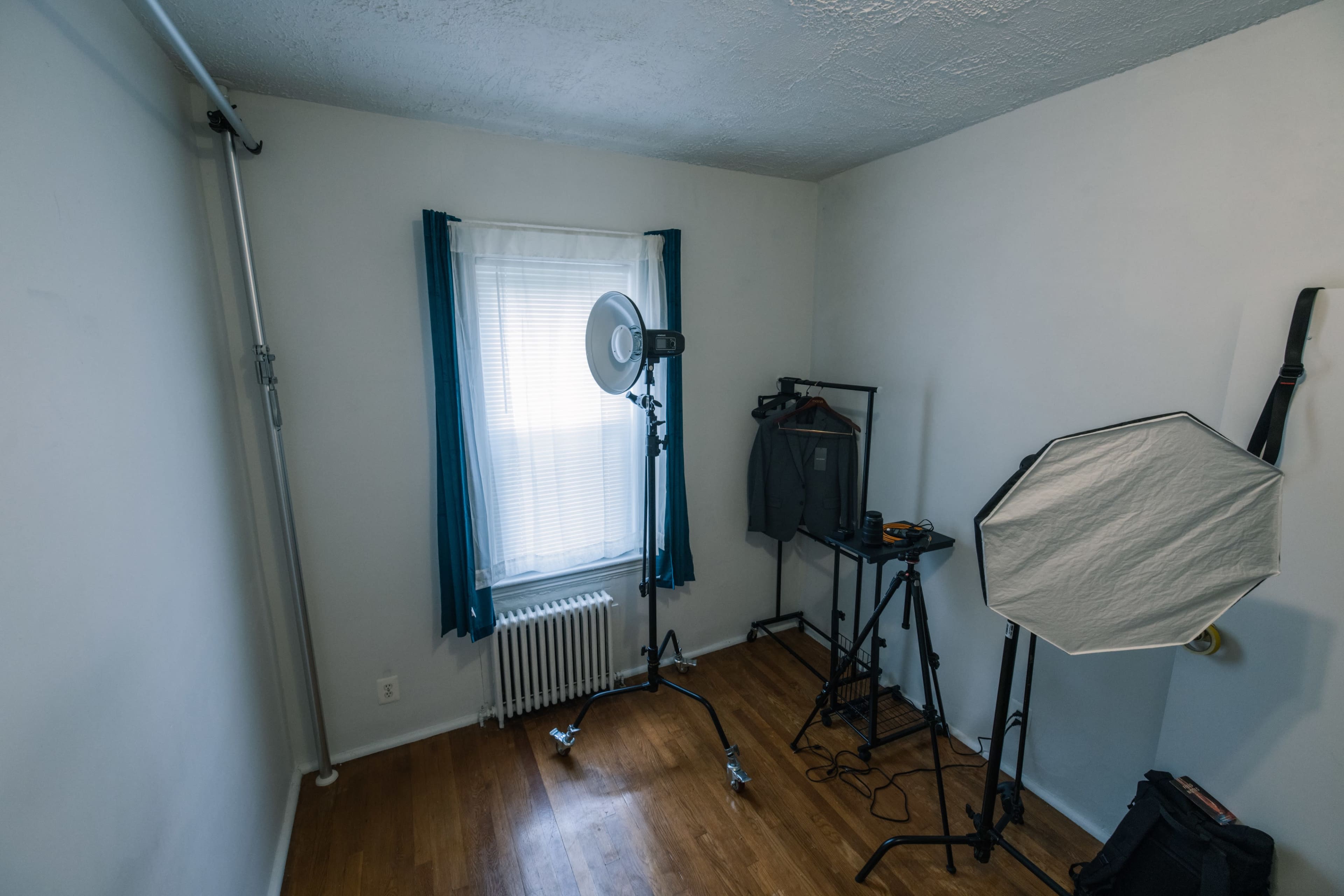 A sparse room with a window, a radiator, and various photography equipment including light stands and a backdrop holder.