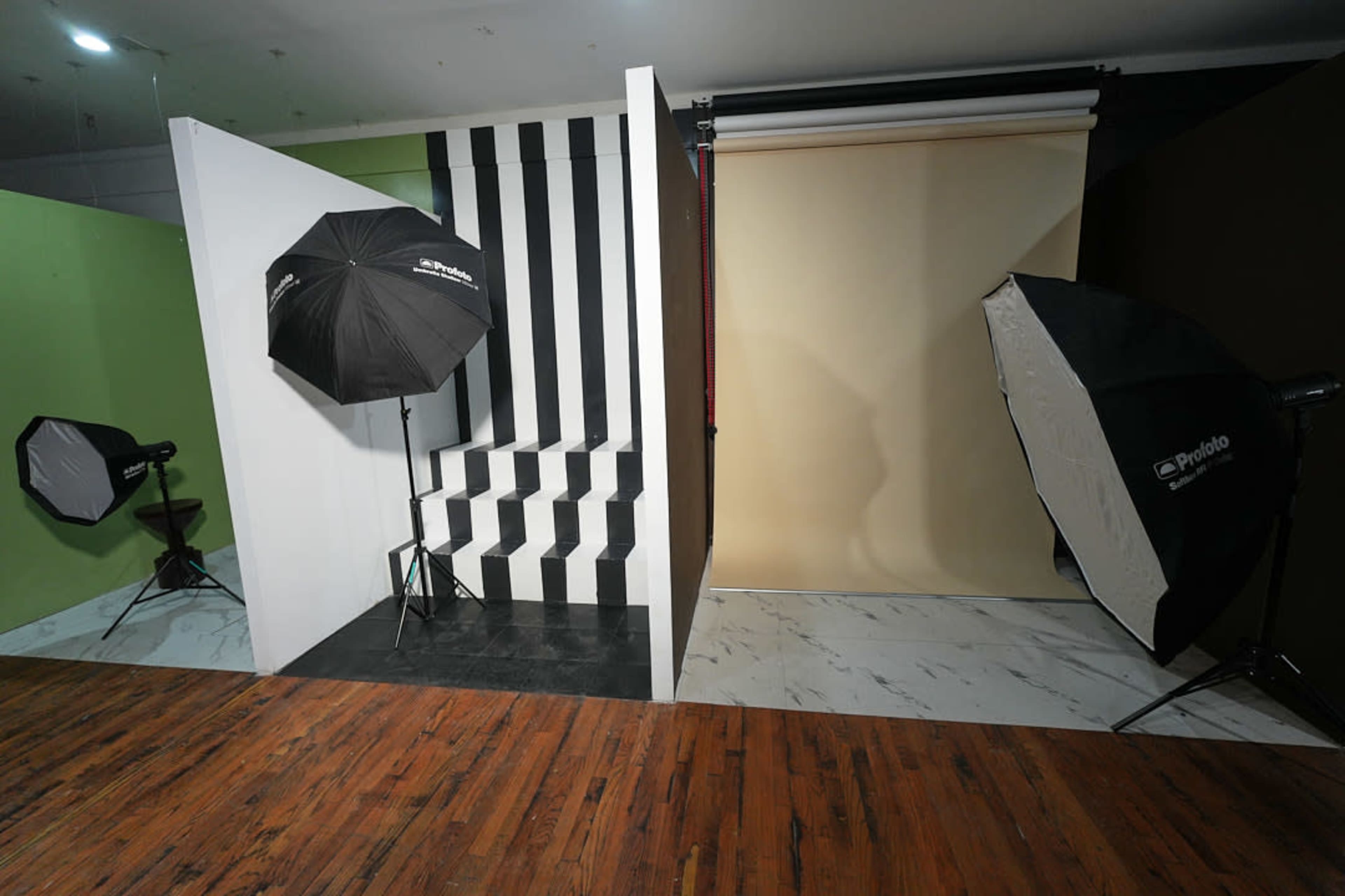 The image shows a photography studio featuring a black and white striped backdrop with stairs, two lighting umbrellas, and a plain brown and beige backdrop section.