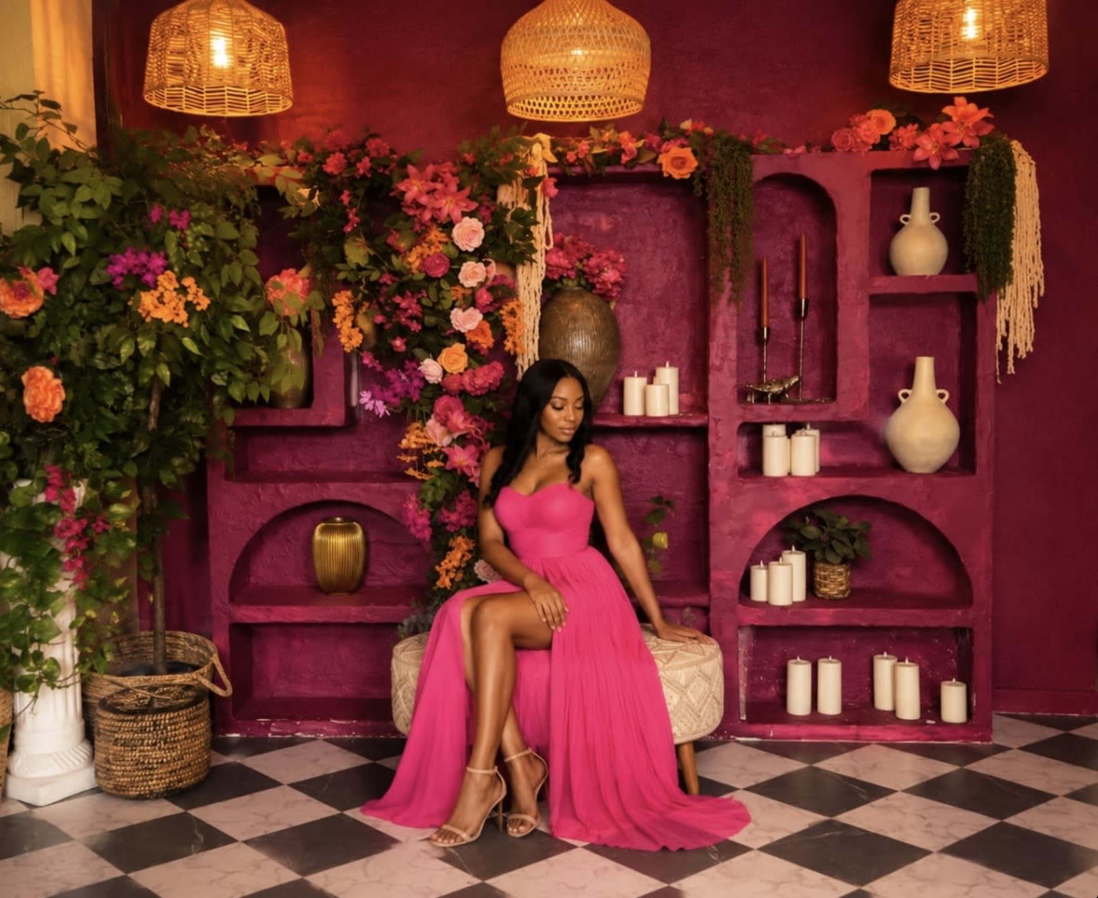 A woman in a pink dress sits on a circular seat surrounded by vibrant flowers and decorative vases against a deep purple wall.