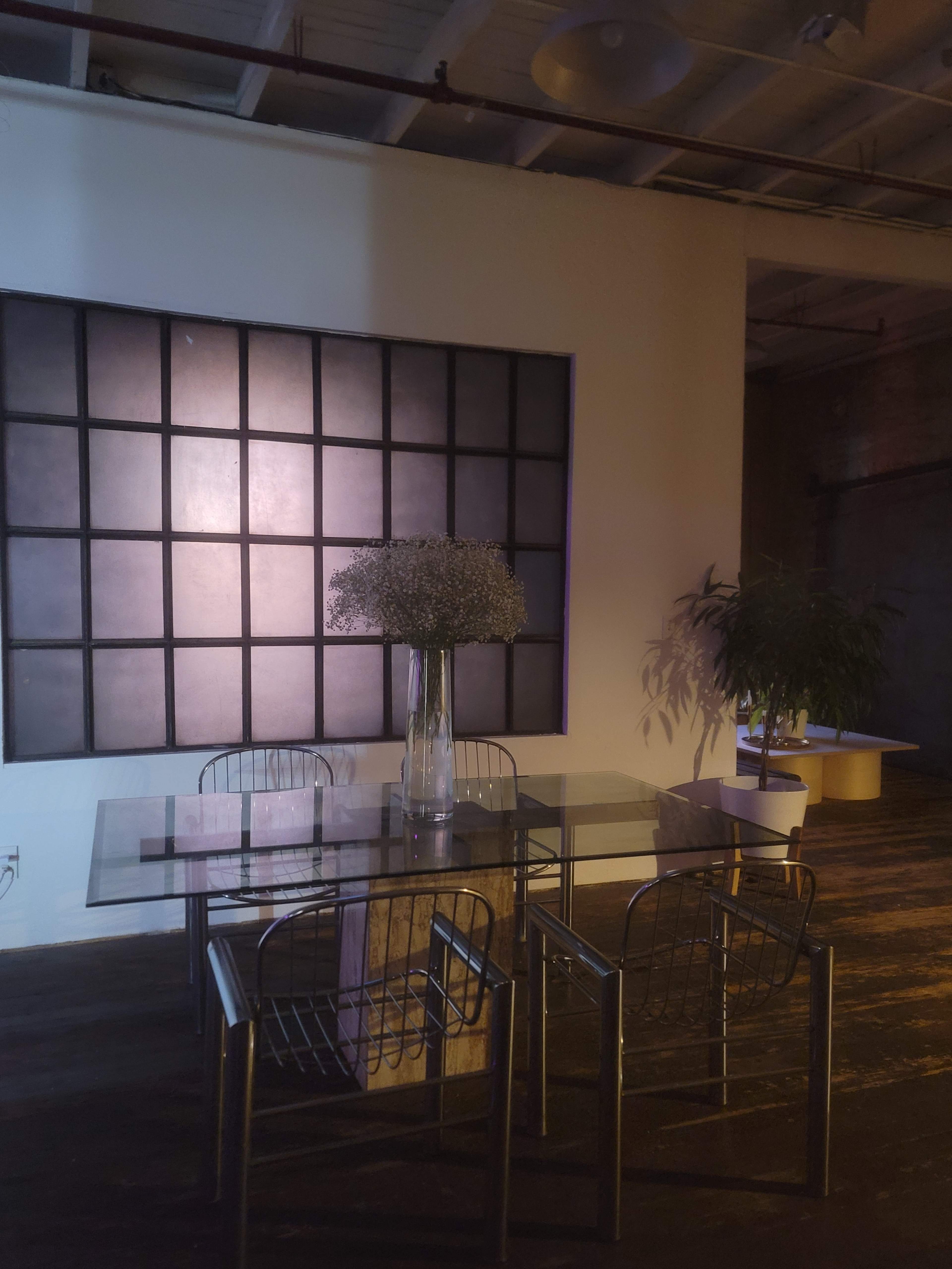 A glass table with metal chairs is situated in a dimly lit room featuring a large grid-patterned wall and a tall vase of dried flowers.