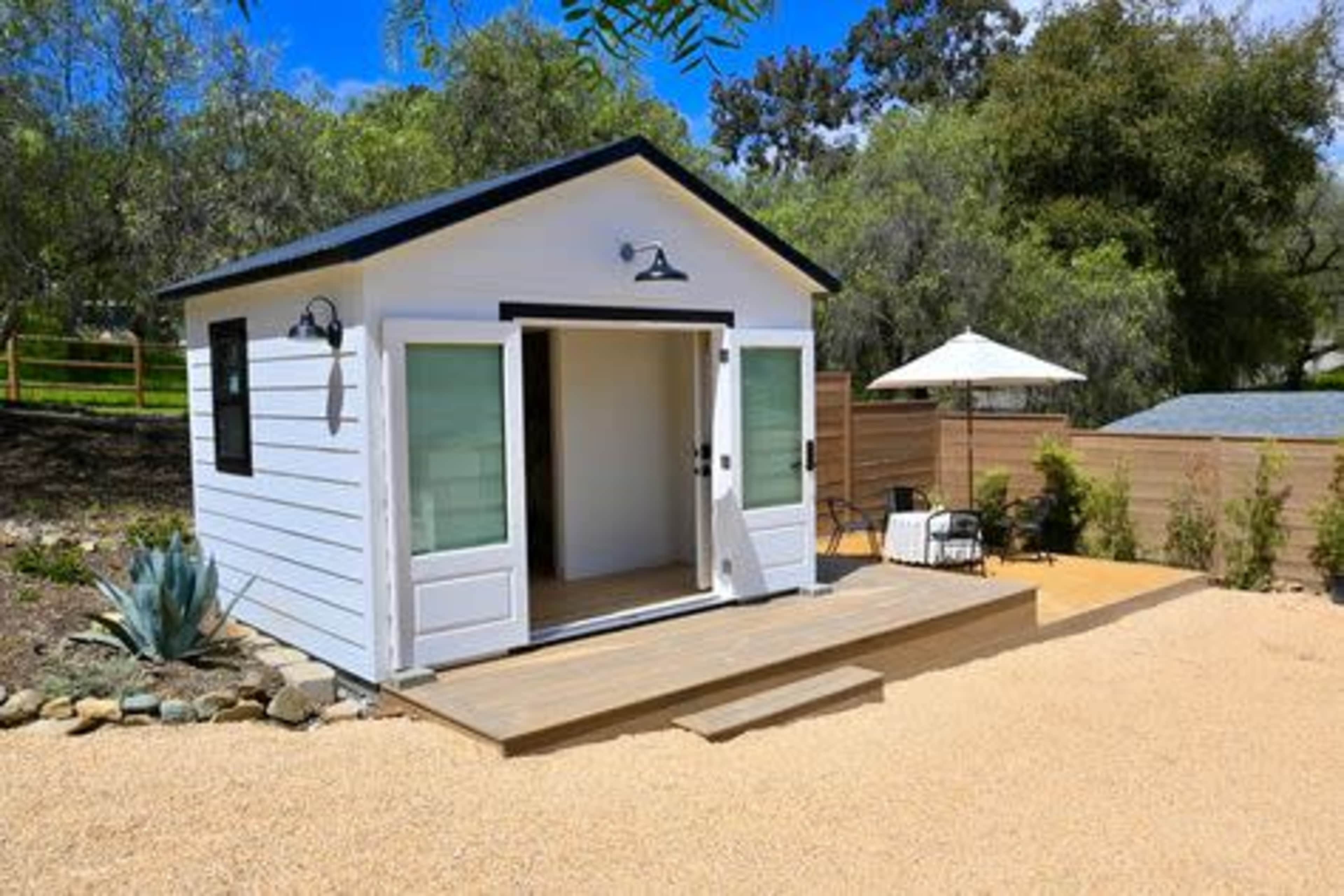 A small modern cabin with a wooden deck and an outdoor seating area is set in a landscaped gravel yard.