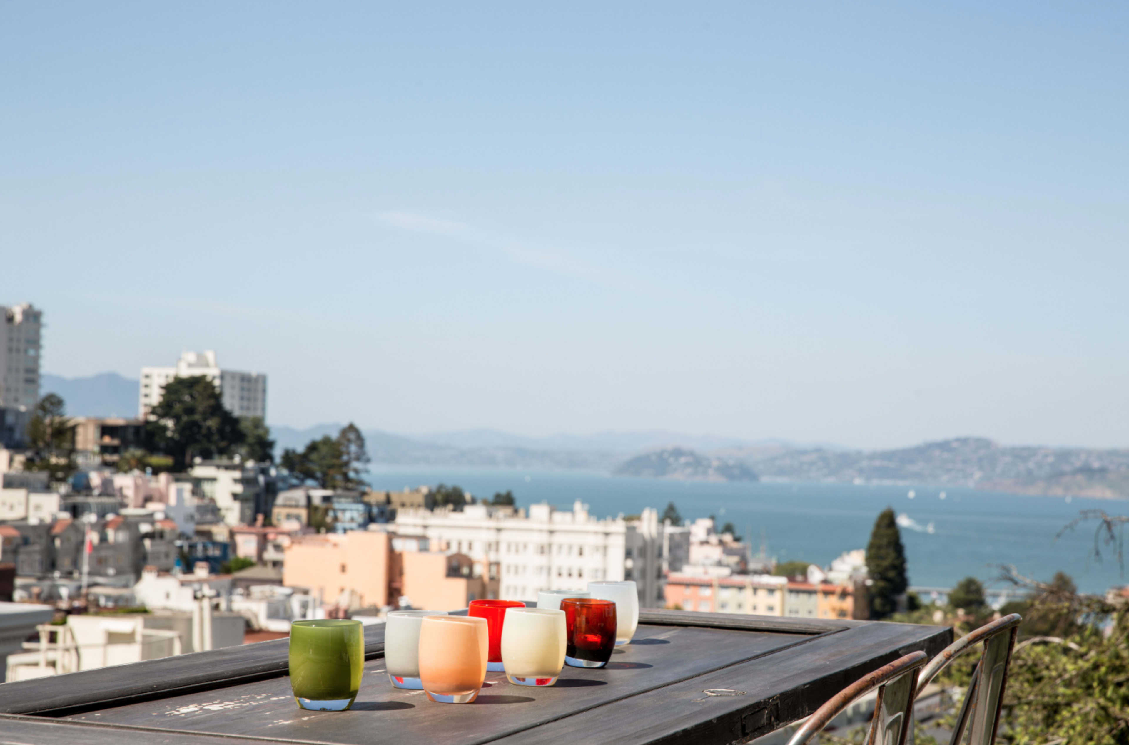 Six colorful glasses are arranged on a wooden table, overlooking a coastal city and body of water in the background.