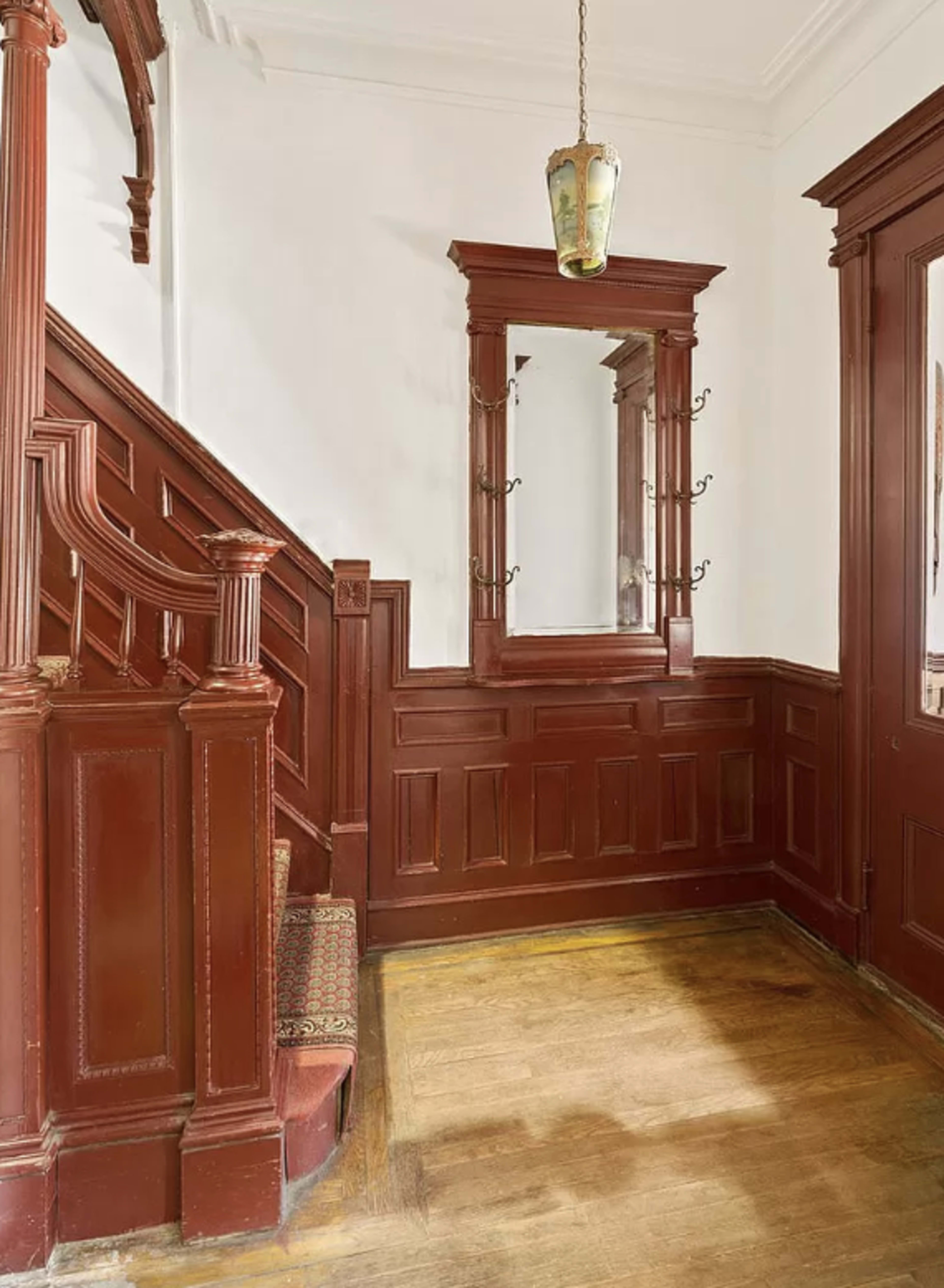 The image shows a wooden staircase with decorative railings leading to a landing, alongside a wall mirror and hooks, all set in a room with dark red wooden paneling.