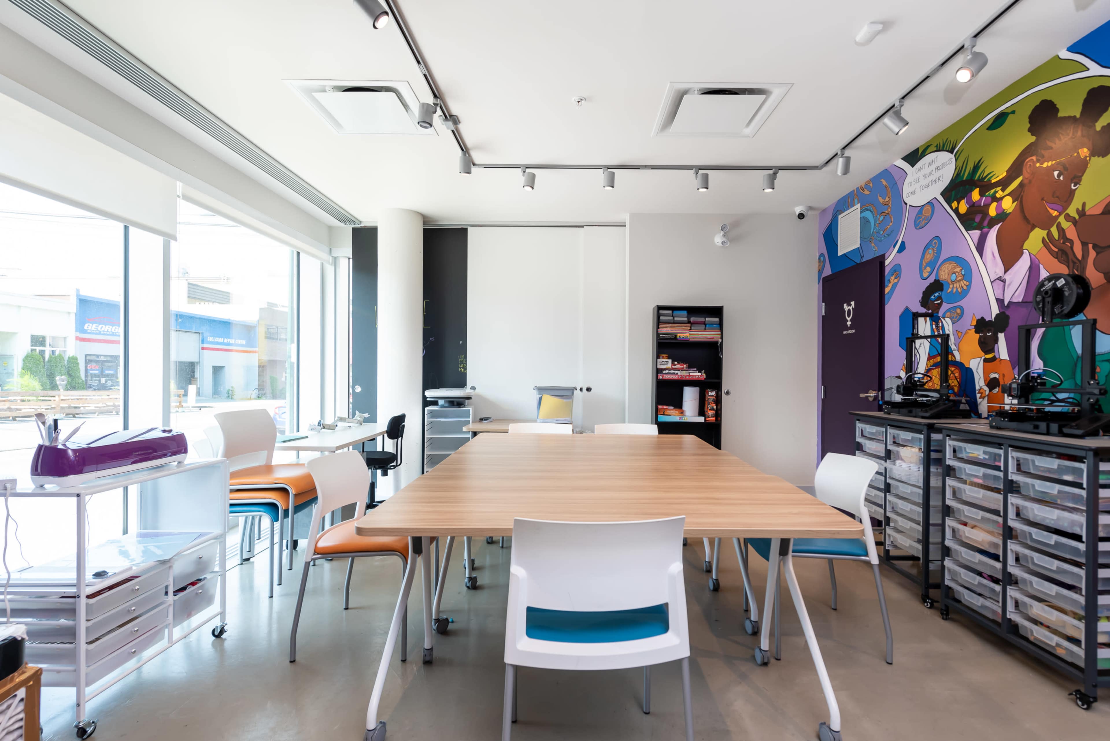 A spacious, modern office features a large wooden table surrounded by colorful chairs, with large windows letting in natural light and a mural on the wall.