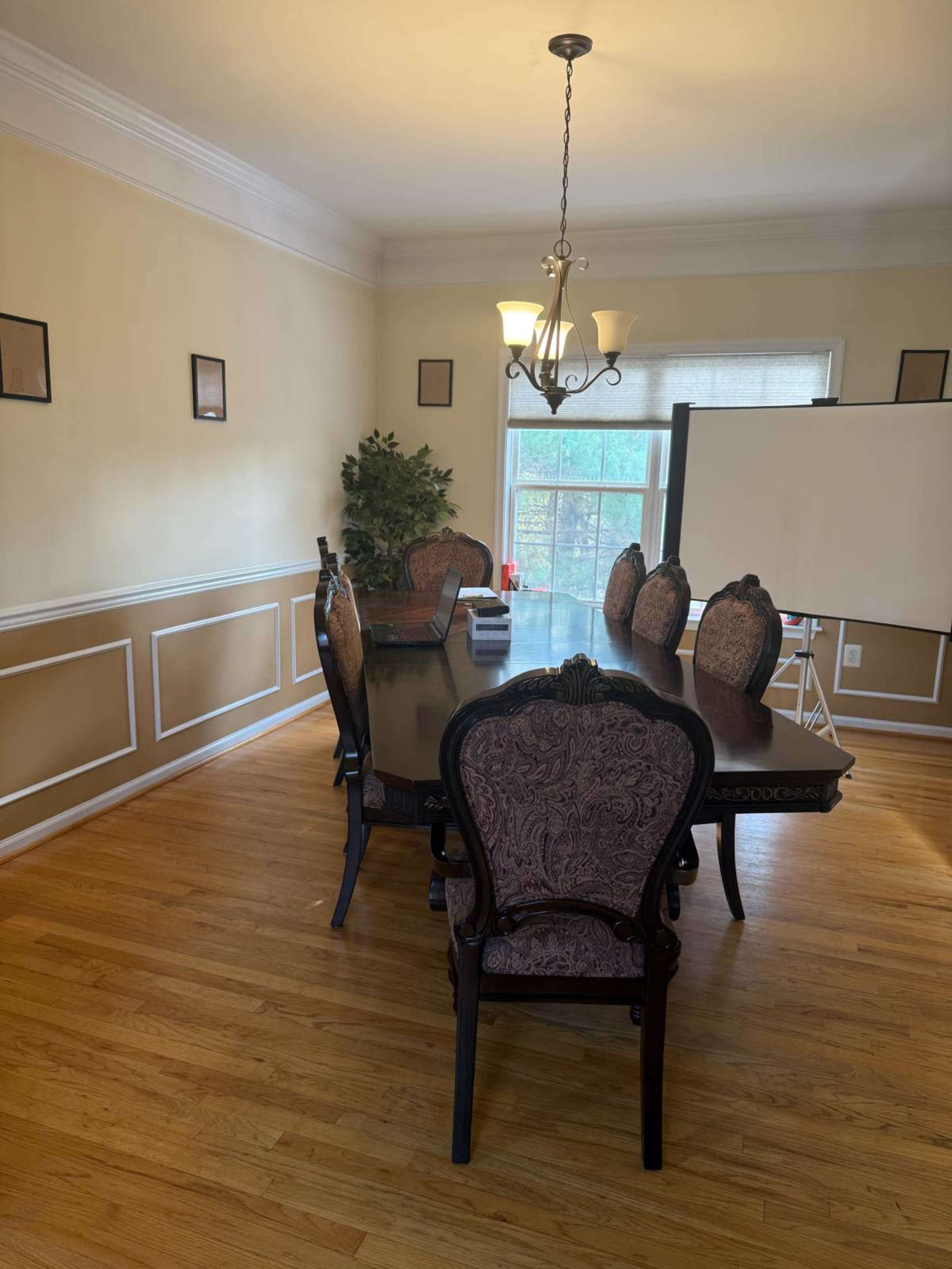 A dining room features a large table surrounded by eight upholstered chairs, with a window and a blank presentation board in the background.