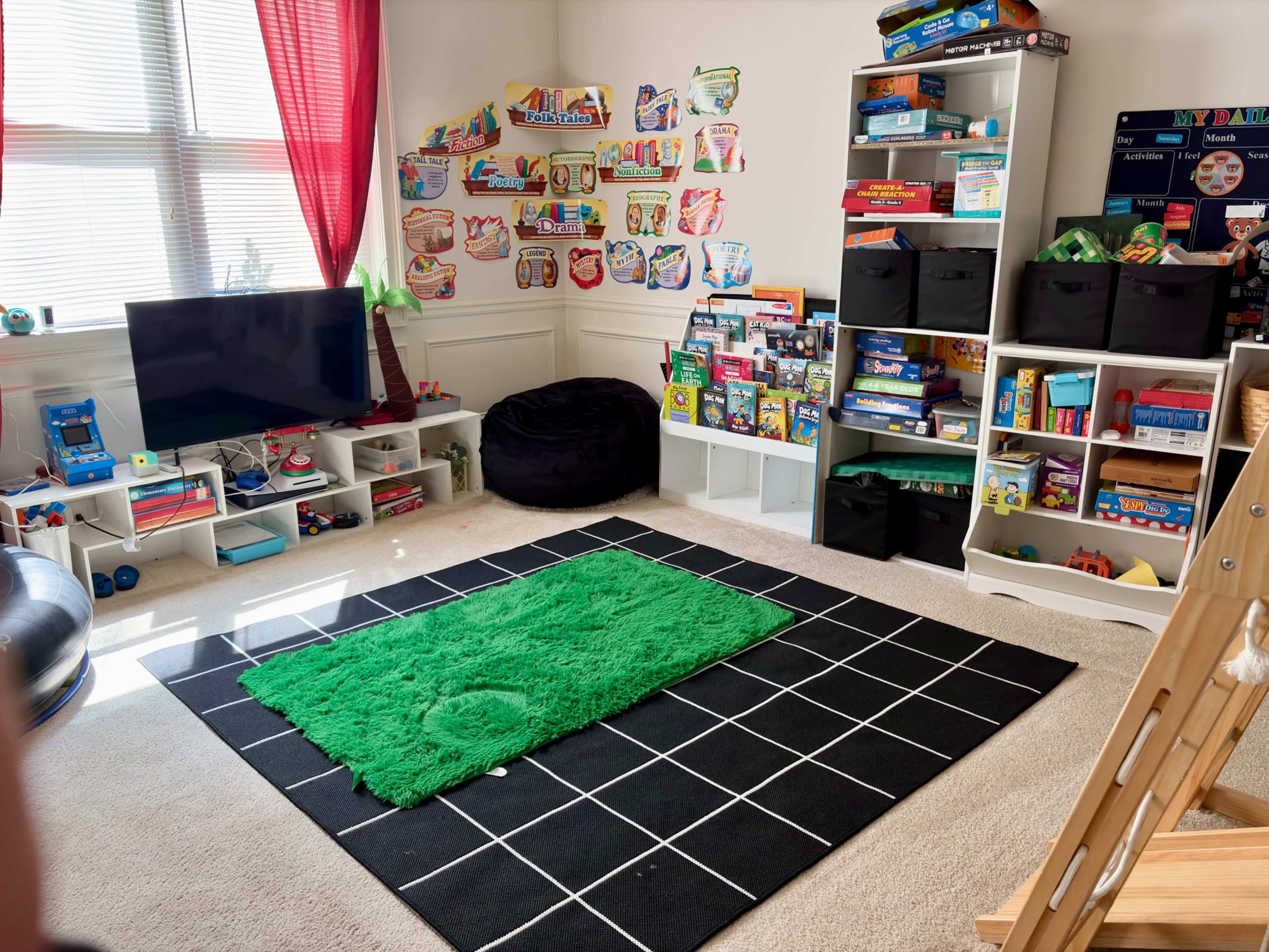 The image shows a playroom with a green rug, a black tiled carpet, a television, and shelves filled with colorful toys and games.