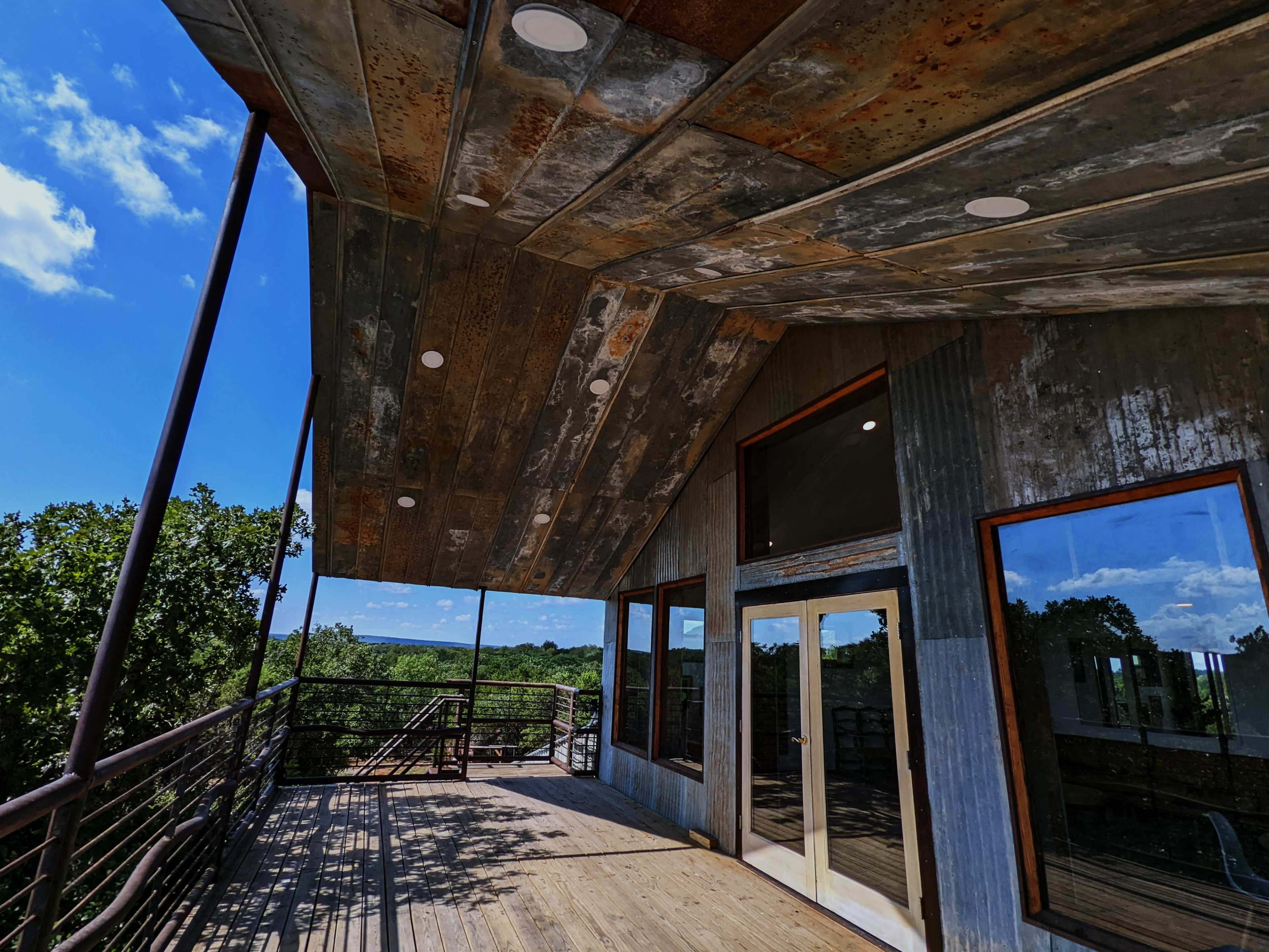 A modern building with a metal roof and wooden deck overlooking a green landscape under a blue sky.
