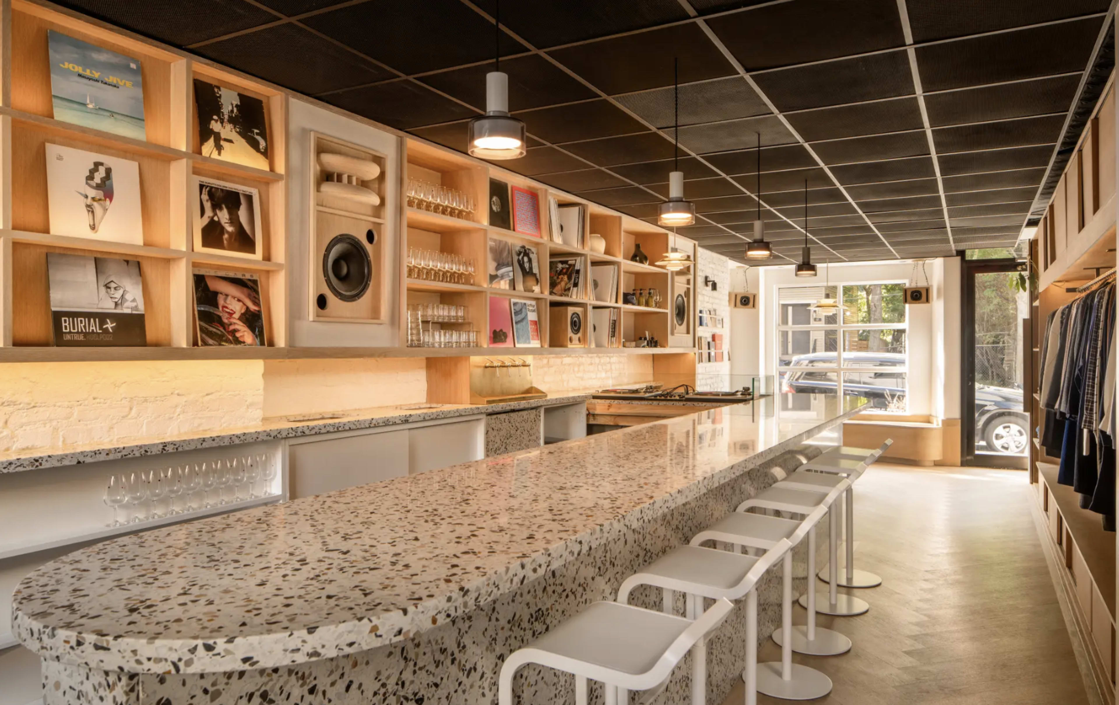 The image shows a modern cafe with a long, speckled countertop, wooden shelves displaying vinyl records, and white stools arranged along the bar area.
