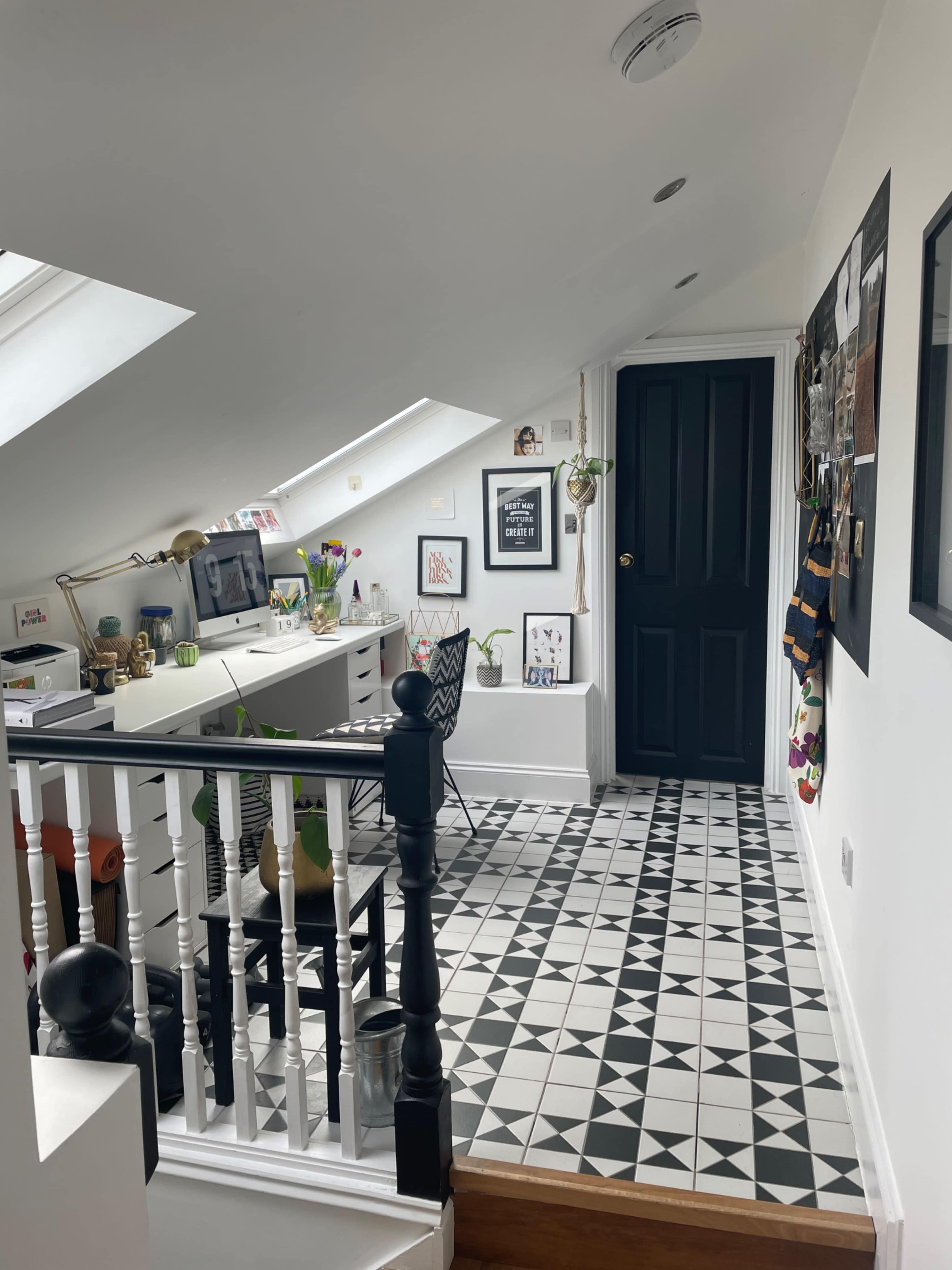 A small room with a sloped ceiling features a desk, decorative items, and a patterned black-and-white tiled floor, leading to a black door.