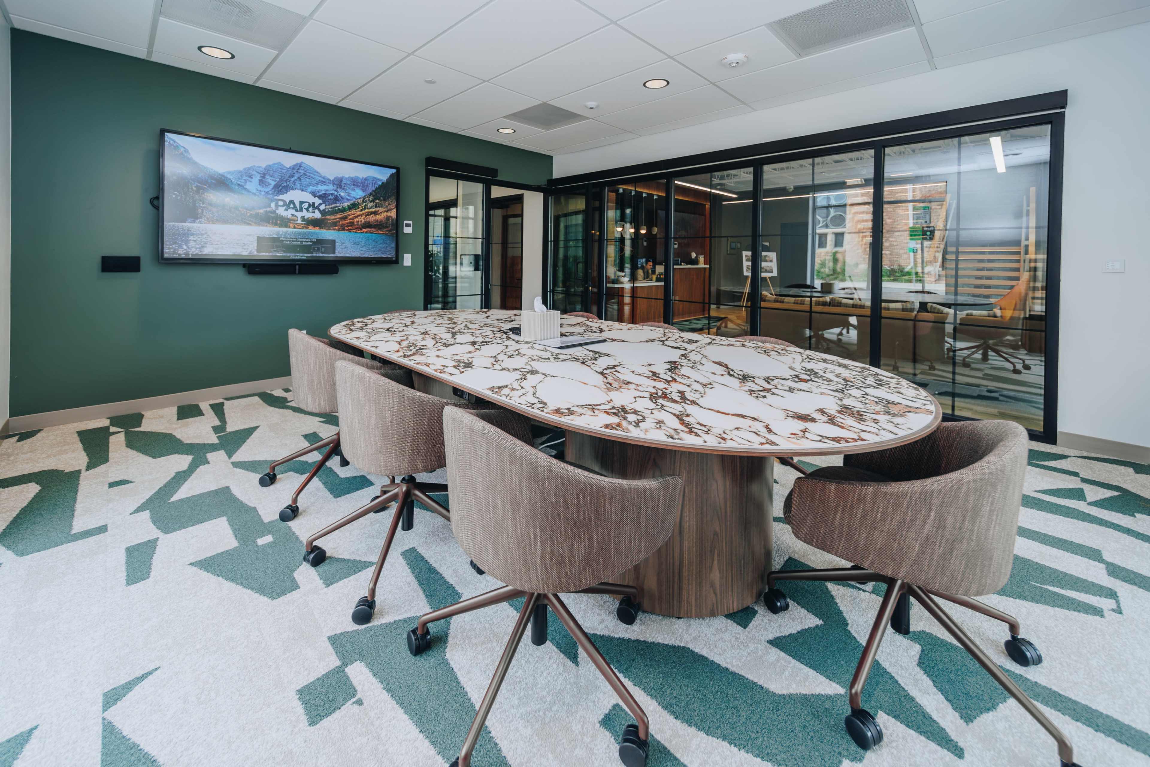 The image shows a modern conference room with a large oval table surrounded by rolling chairs, a wall-mounted TV, and glass panels separating it from another area.