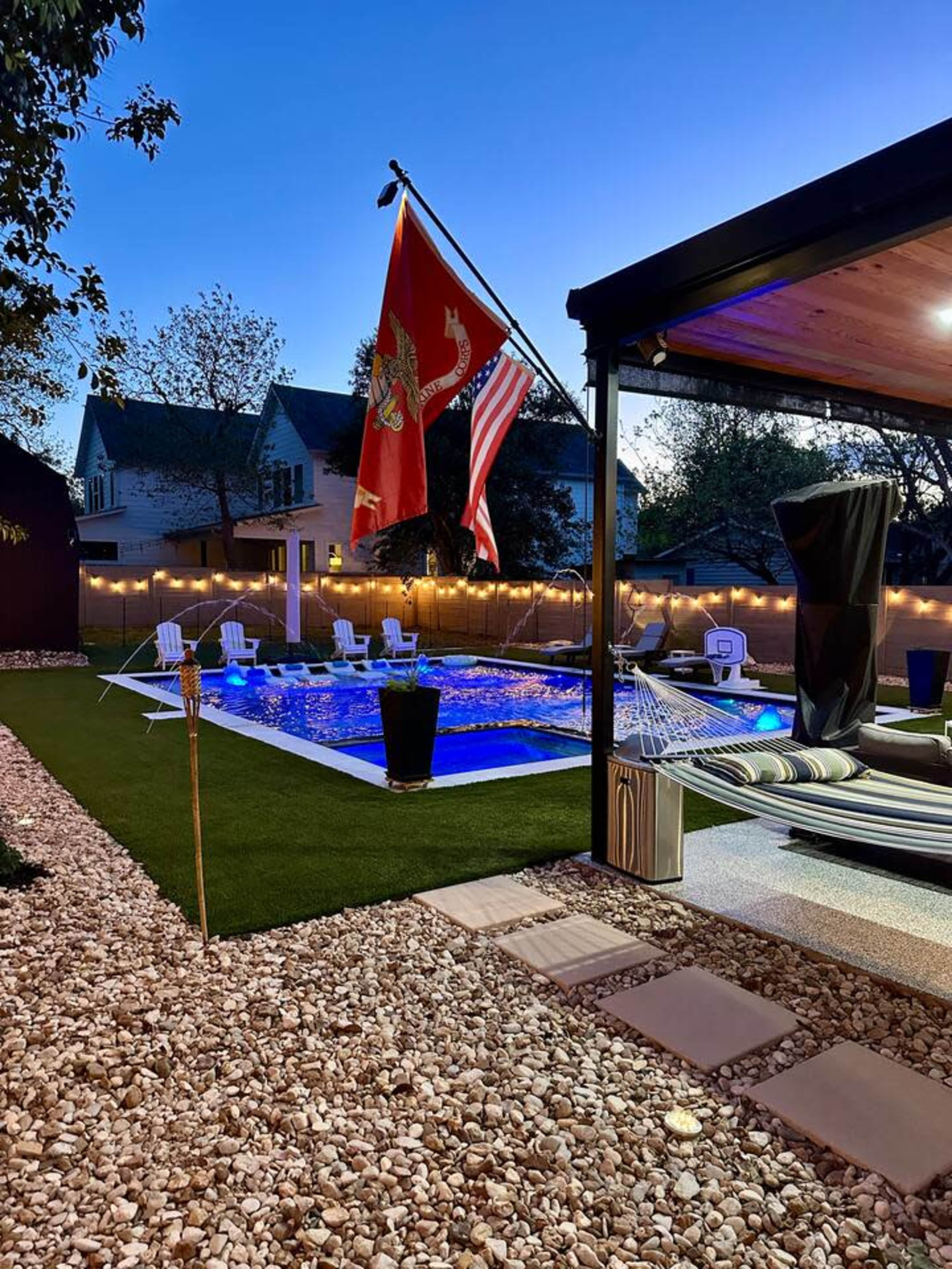 The image shows a well-lit backyard with a swimming pool, surrounded by decorative stones, chairs, and two flags flying near the pool.