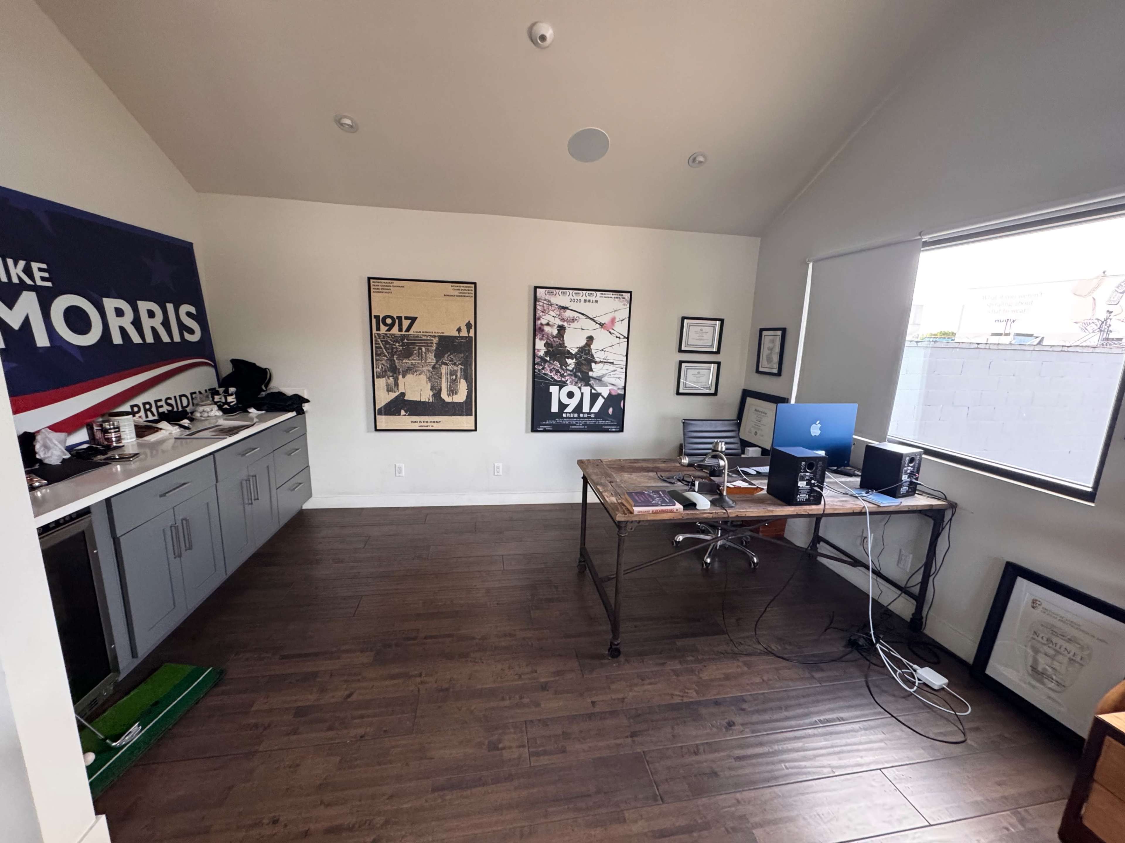 The image shows a modern office space with a wooden desk, a computer, and two framed posters on the walls, along with a large banner.