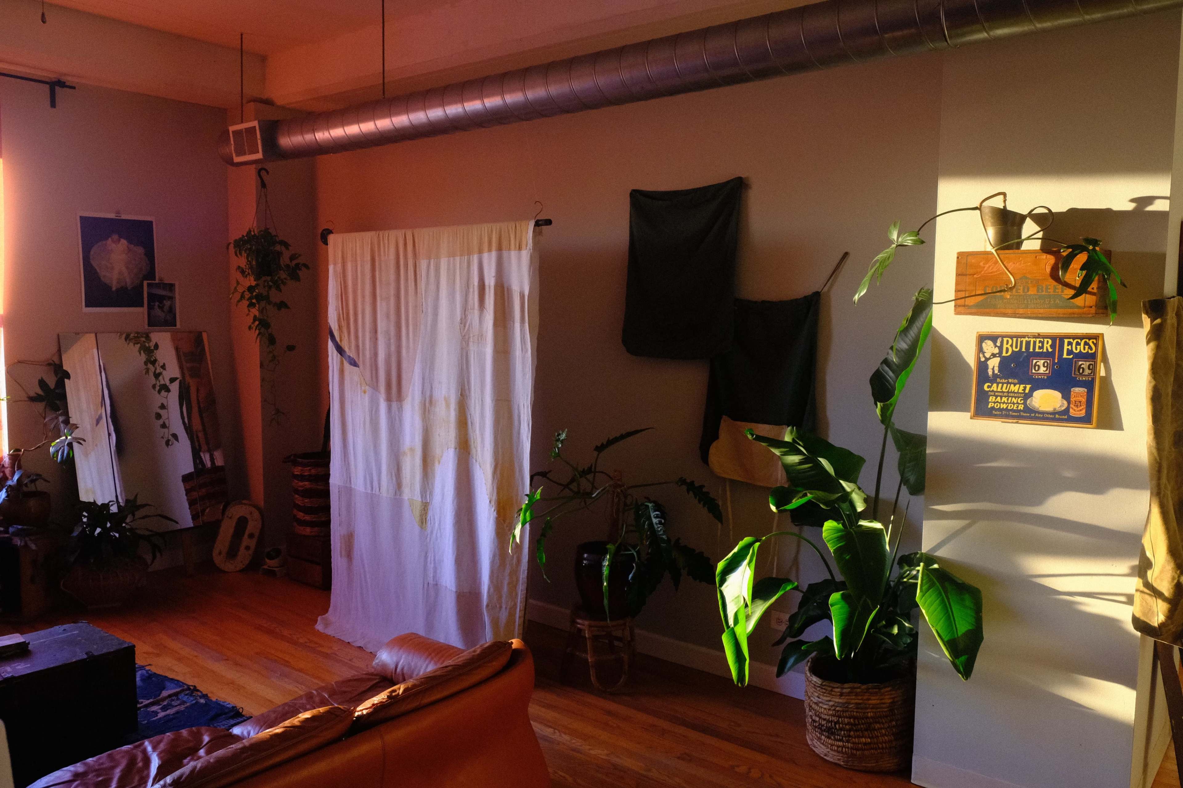The image shows a cozy room with a leather sofa, plants in woven baskets, a sheer curtain dividing the space, and various textiles hanging on the walls.