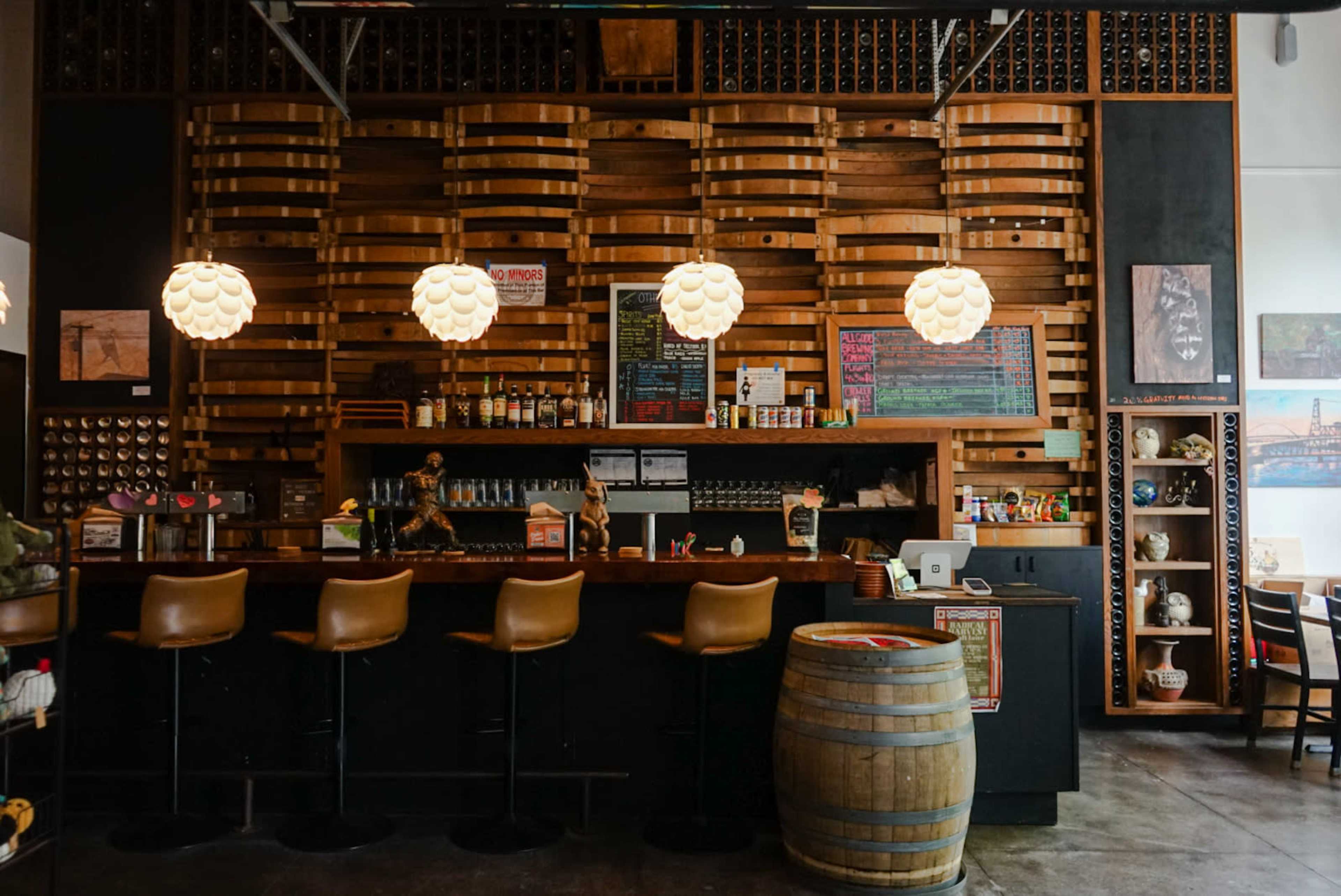 The image shows a cozy bar area featuring wooden shelves, three pendant lights, and a counter with high stools and a barrel.