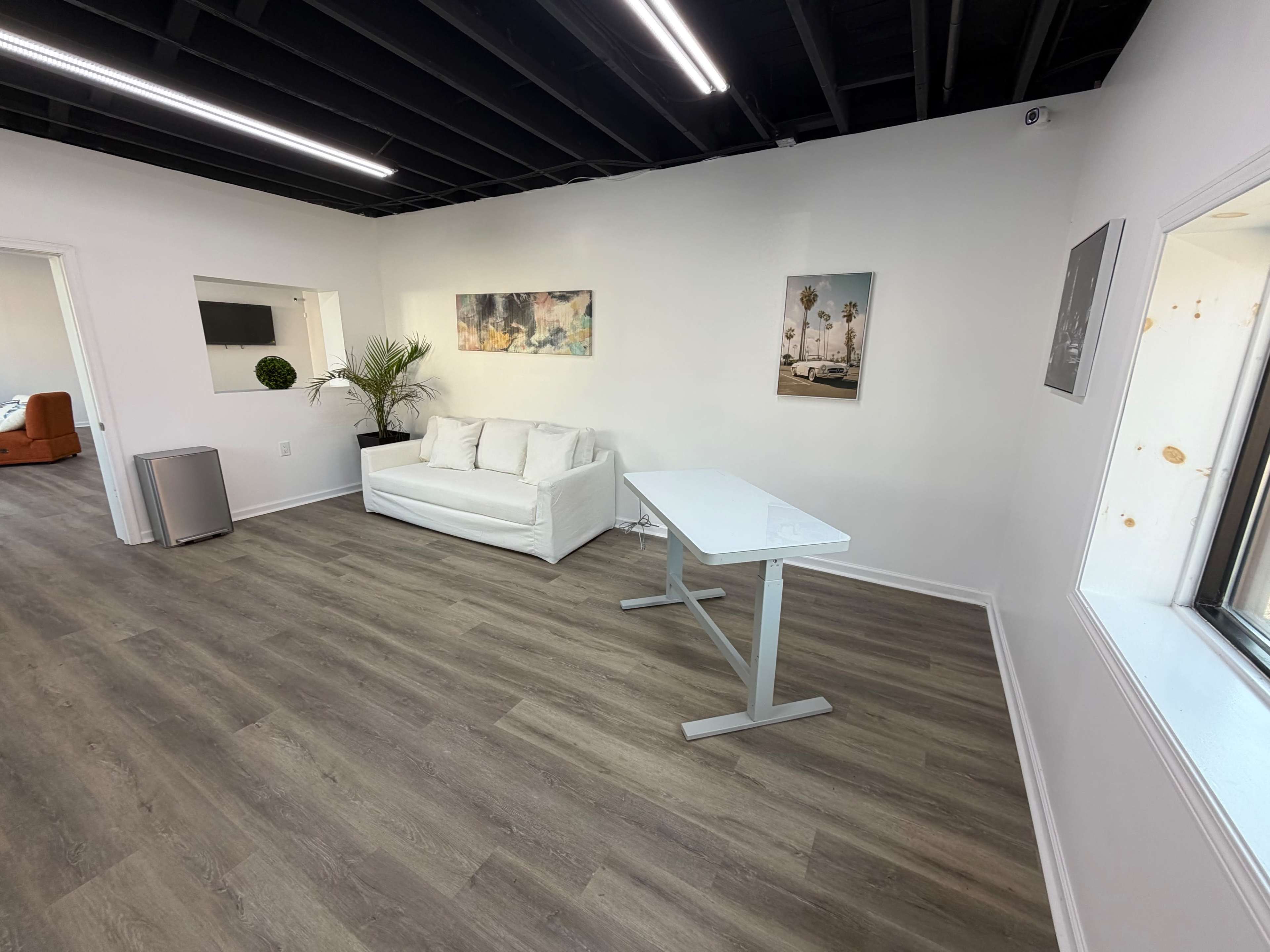The image shows a modern, minimalistic office space with a white couch, a glass table, a plant in the corner, and framed pictures on the walls.