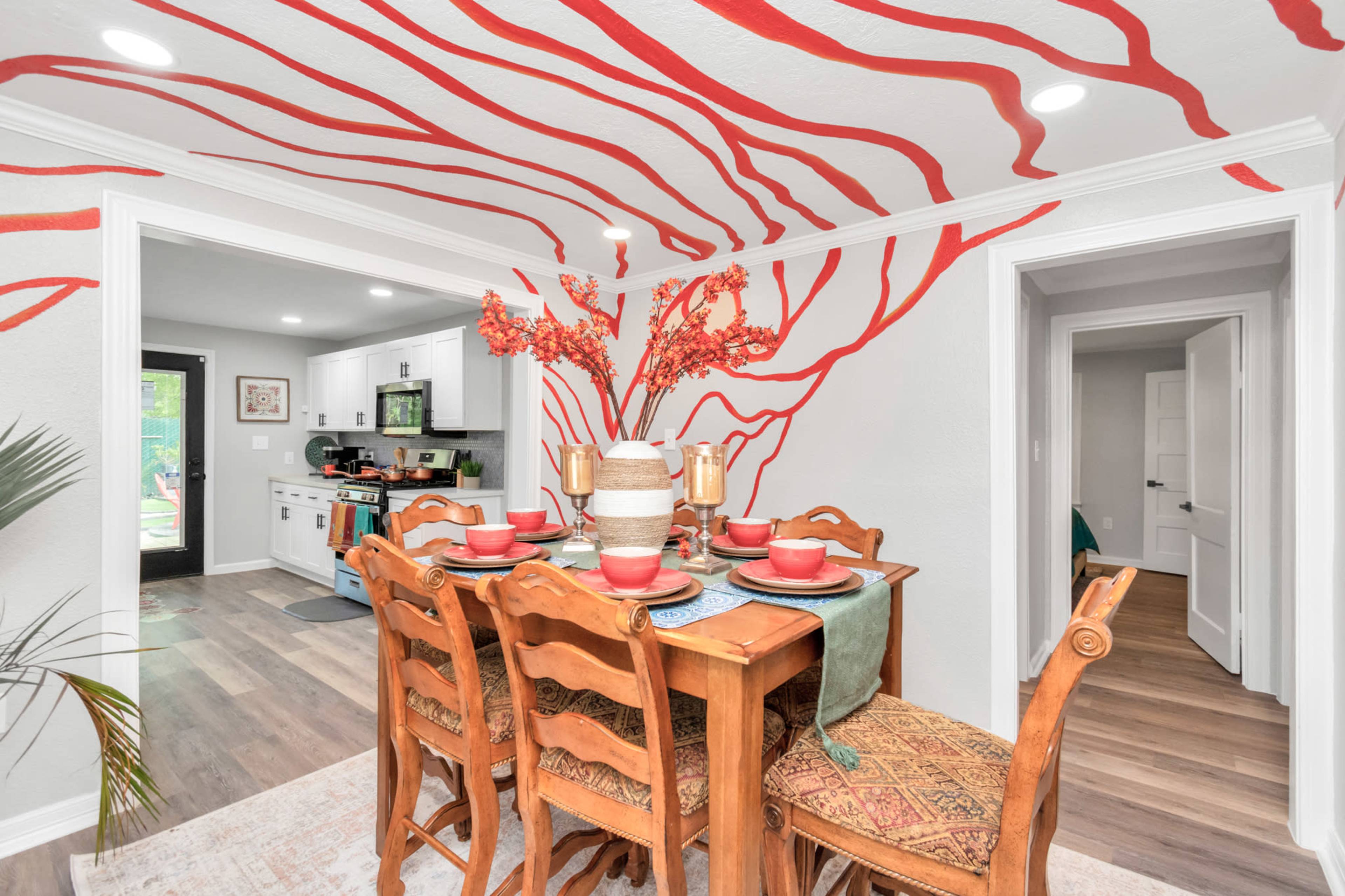 A dining area features a wooden table set for six, with red plates and a large vase of flowers, against a backdrop of bold red lines on the wall.