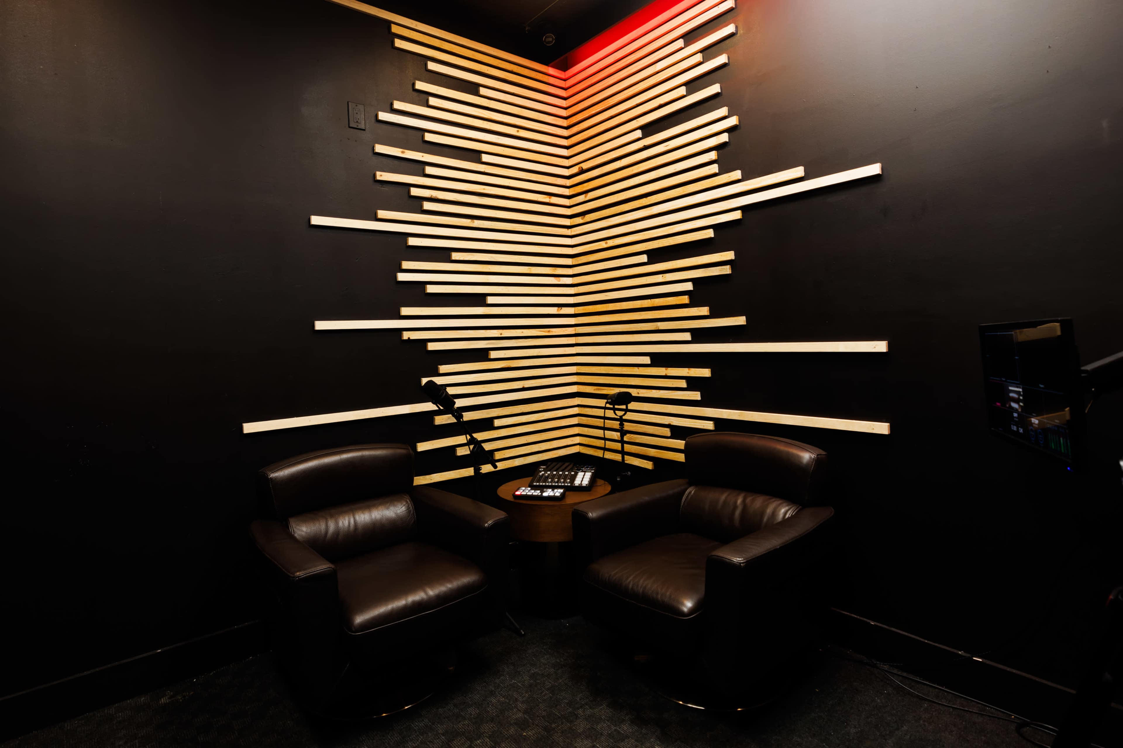 The image depicts a corner of a room with two leather chairs facing a small table, surrounded by a wall featuring a geometric arrangement of wooden strips.
