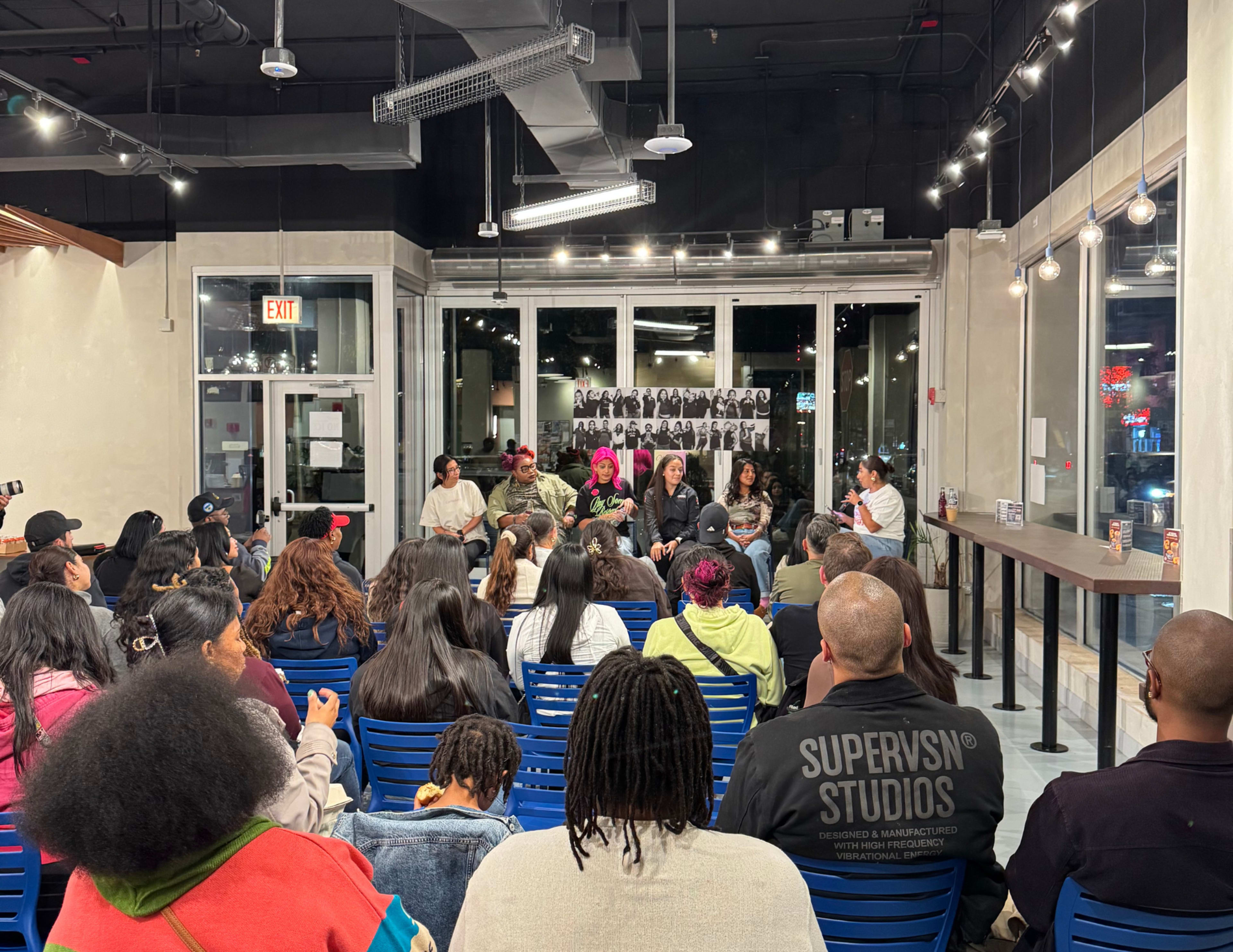 A diverse group of people sits in rows of blue chairs facing a panel discussion inside a well-lit café.