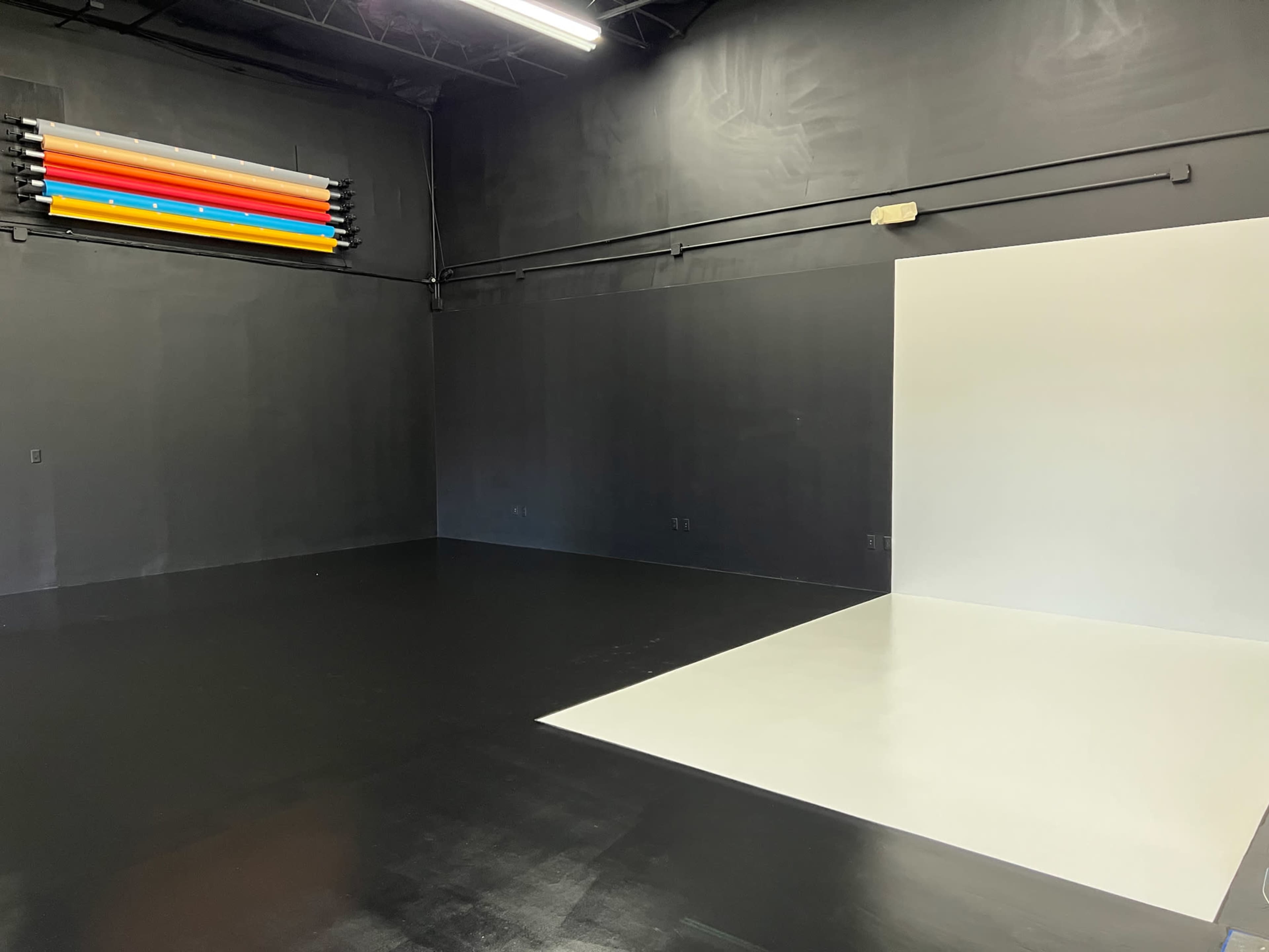 An empty studio space with a black floor, dark walls, and a white backdrop positioned on one side.