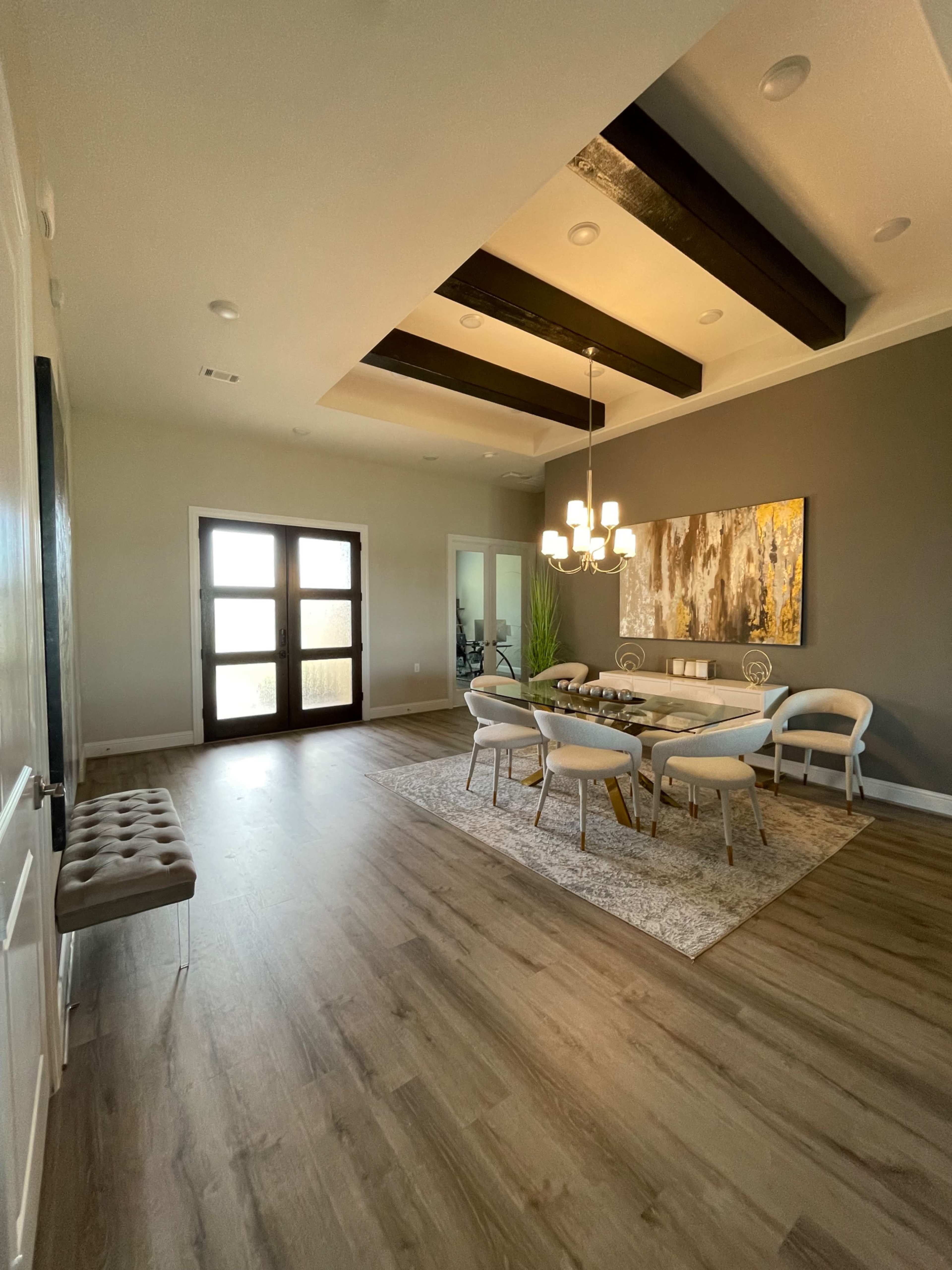 A dining area features a round table surrounded by chairs, with large windows and a coffered ceiling illuminated by a modern chandelier.