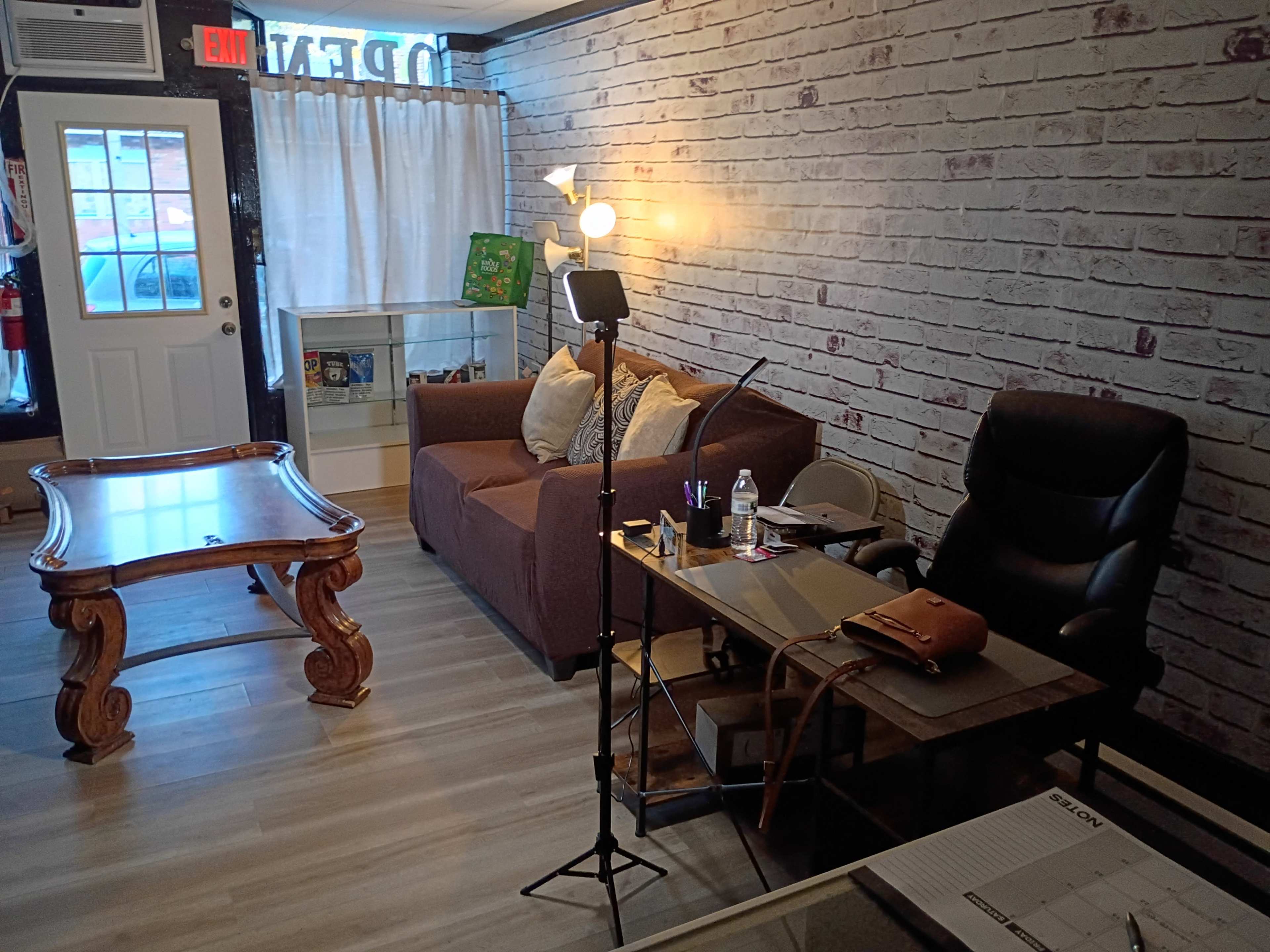 The image shows a cozy indoor space with a brown sofa, a wooden coffee table, a desk with a chair, and a floor lamp, all against a wall with a brick pattern.