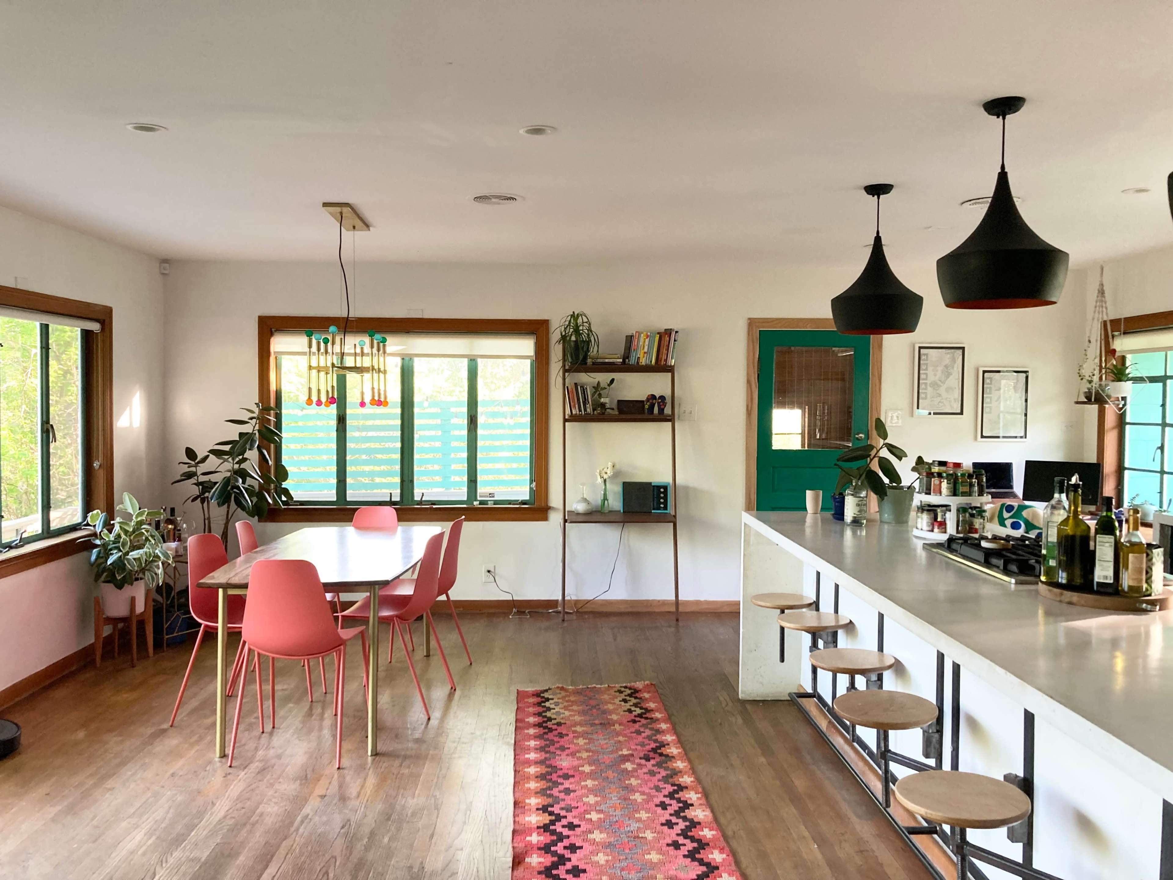 A modern kitchen and dining area featuring a round table with pink chairs, a long bar with stools, and large windows allowing natural light.