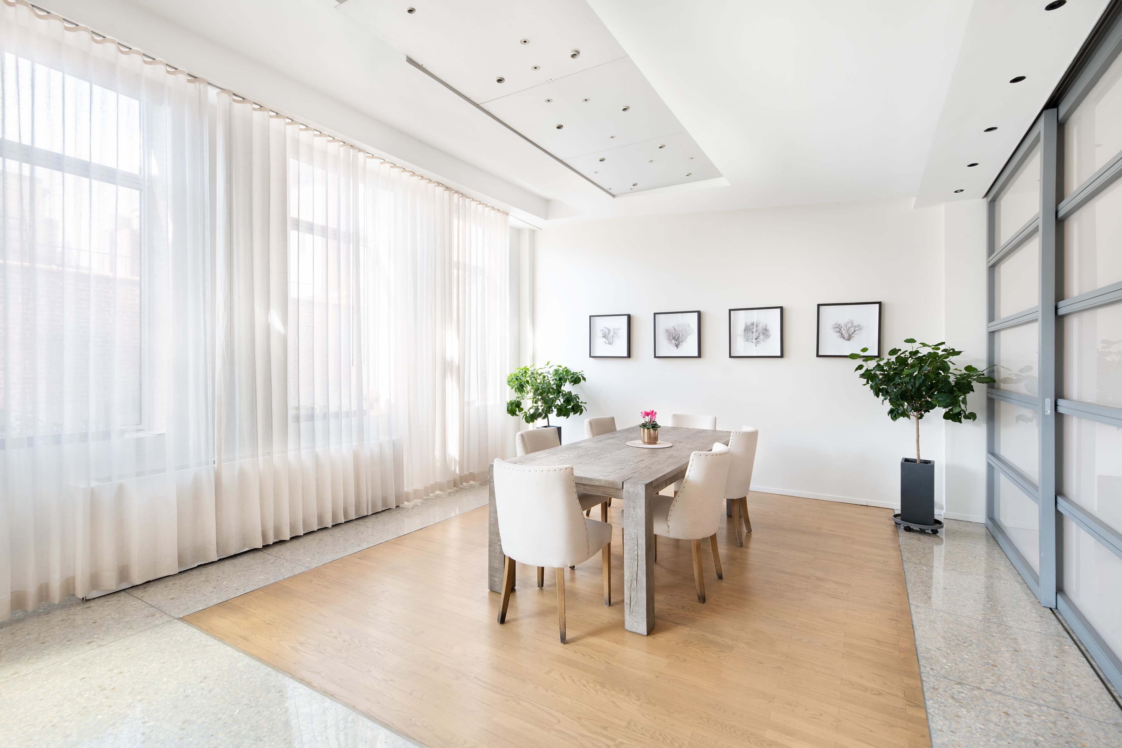 A dining area features a wooden table surrounded by six chairs, with large windows draped in sheer curtains and two plants in the corners.