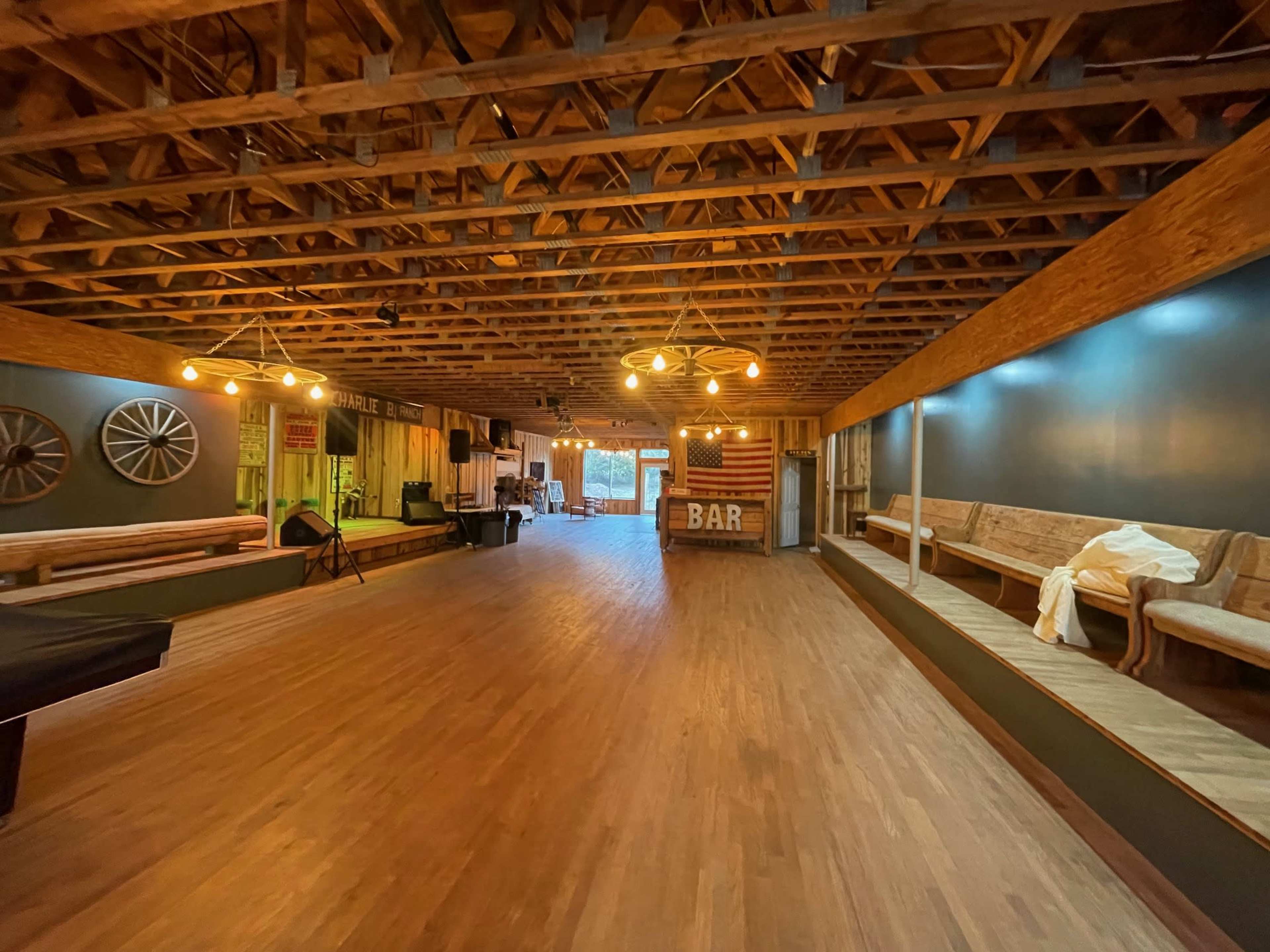 A spacious interior with wooden flooring, exposed beams, and a bar area in the corner, illuminated by hanging light fixtures.