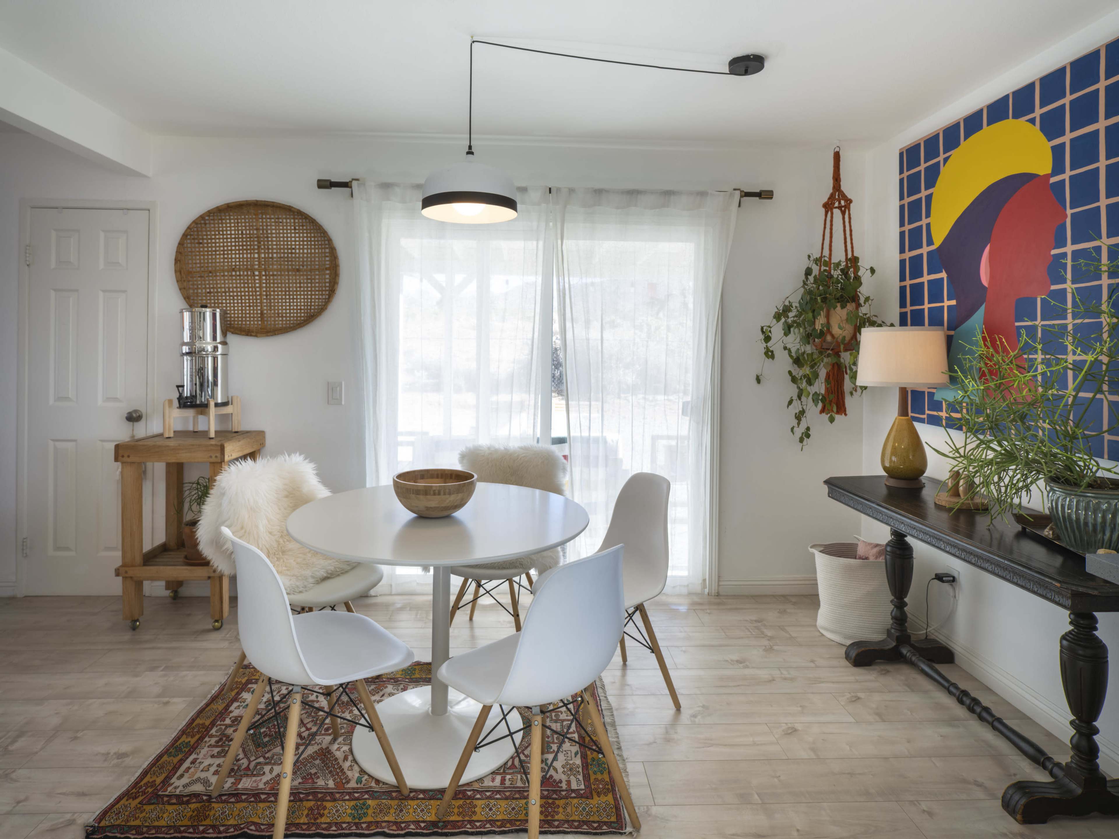 A modern dining area features a round table with white chairs, a patterned rug, and a large wall art piece against a bright window backdrop.