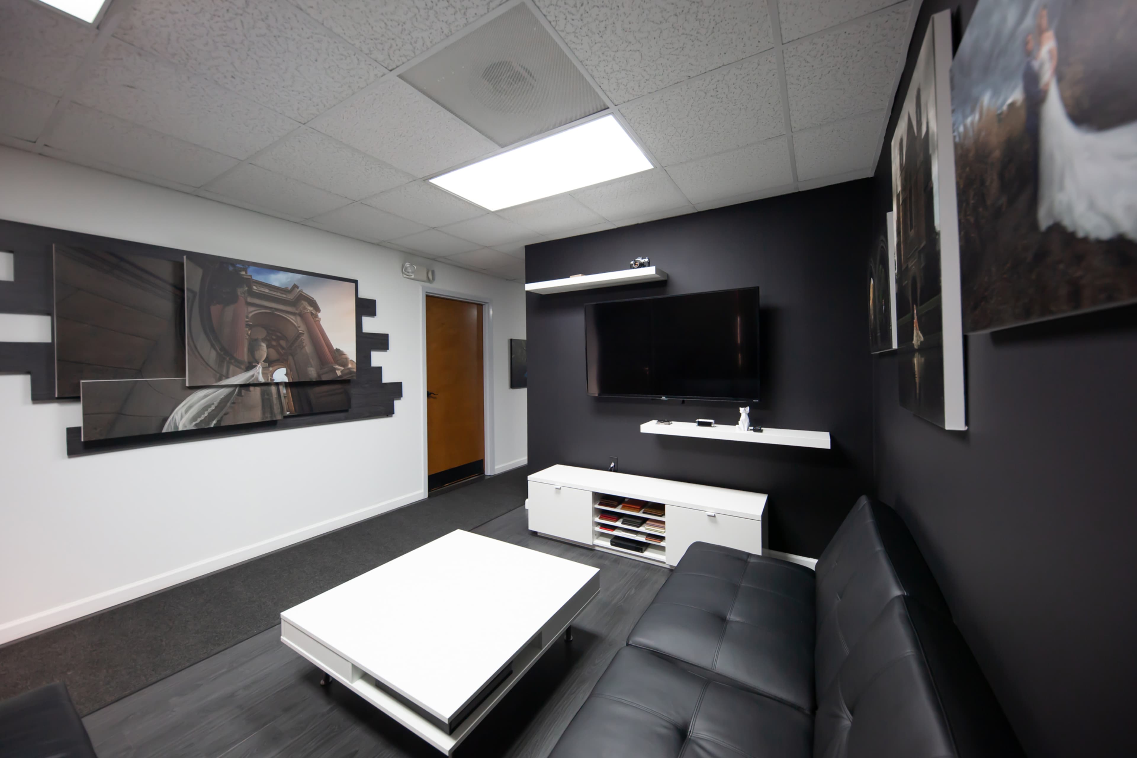 A modern lounge area features a black sofa, a white coffee table, and a wall-mounted TV, with art pieces and a door leading to another room.