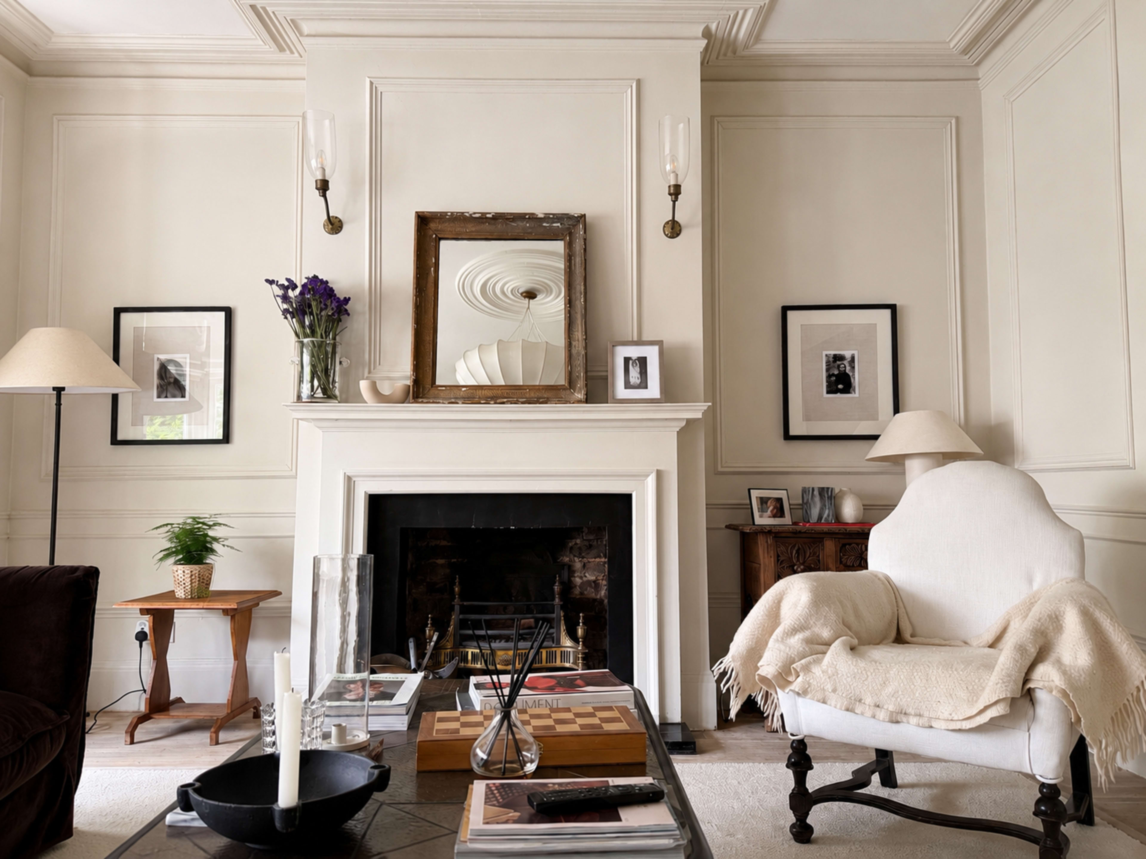 The image shows a cozy living room with a white fireplace, a large mirror above it, framed photographs on the walls, and a comfortable armchair draped with a light blanket.
