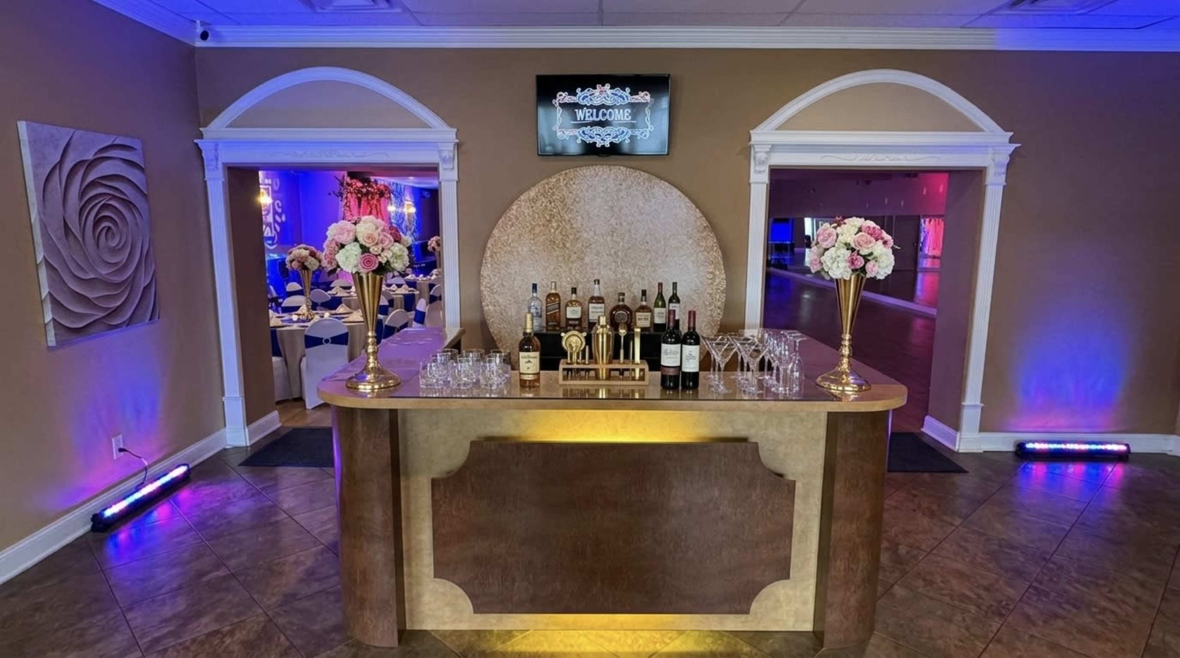 The image shows a decorated bar area in an event space, featuring floral arrangements and a display of liquor bottles alongside glasses.