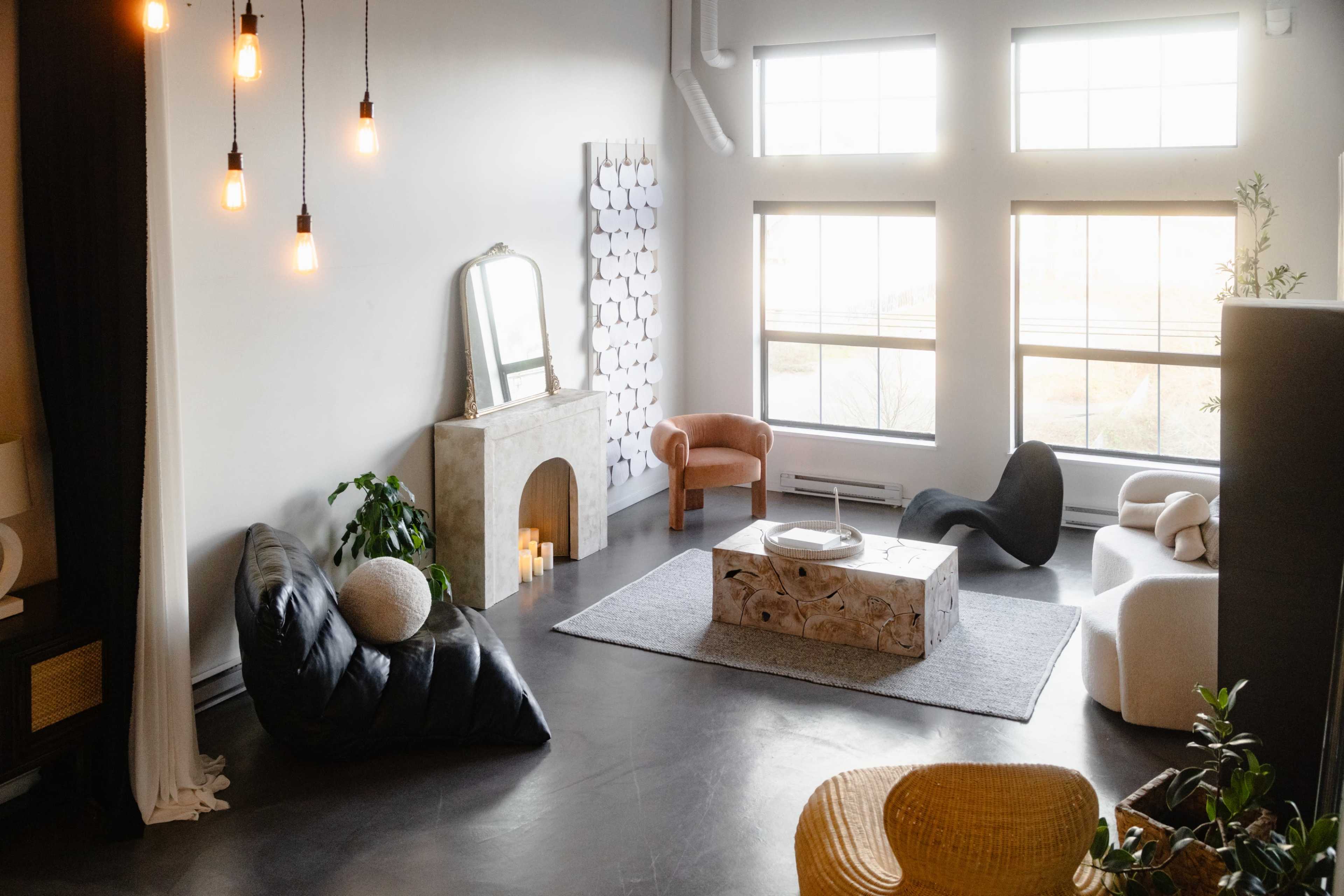 The image shows a modern, spacious living area featuring a variety of furniture pieces, including a black leather sofa, a sculptural coffee table, and a light-colored armchair, with large windows allowing natural light to fill the room.