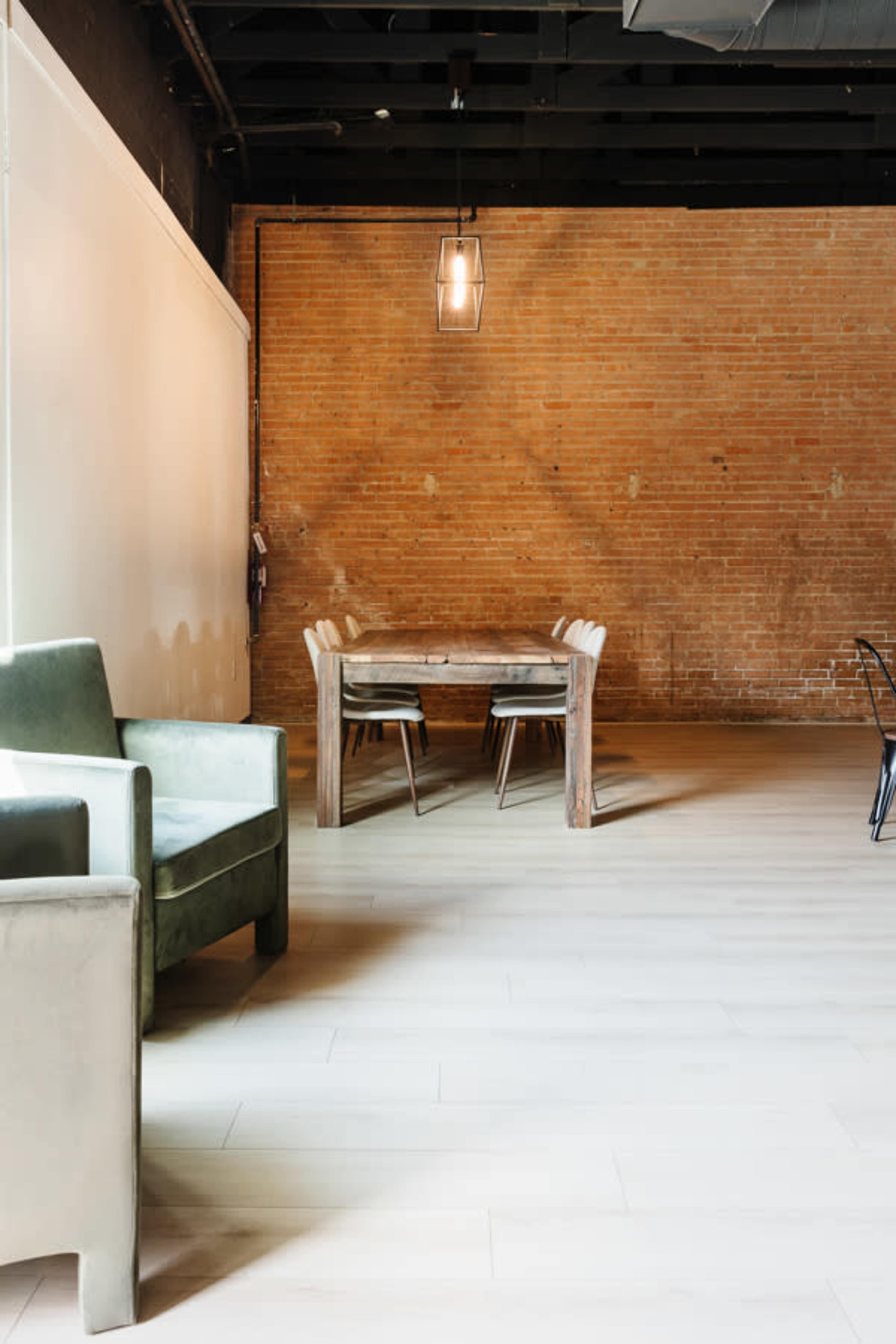 A minimalist interior with a wooden table and chairs against a textured brick wall, complemented by a green armchair in the foreground.
