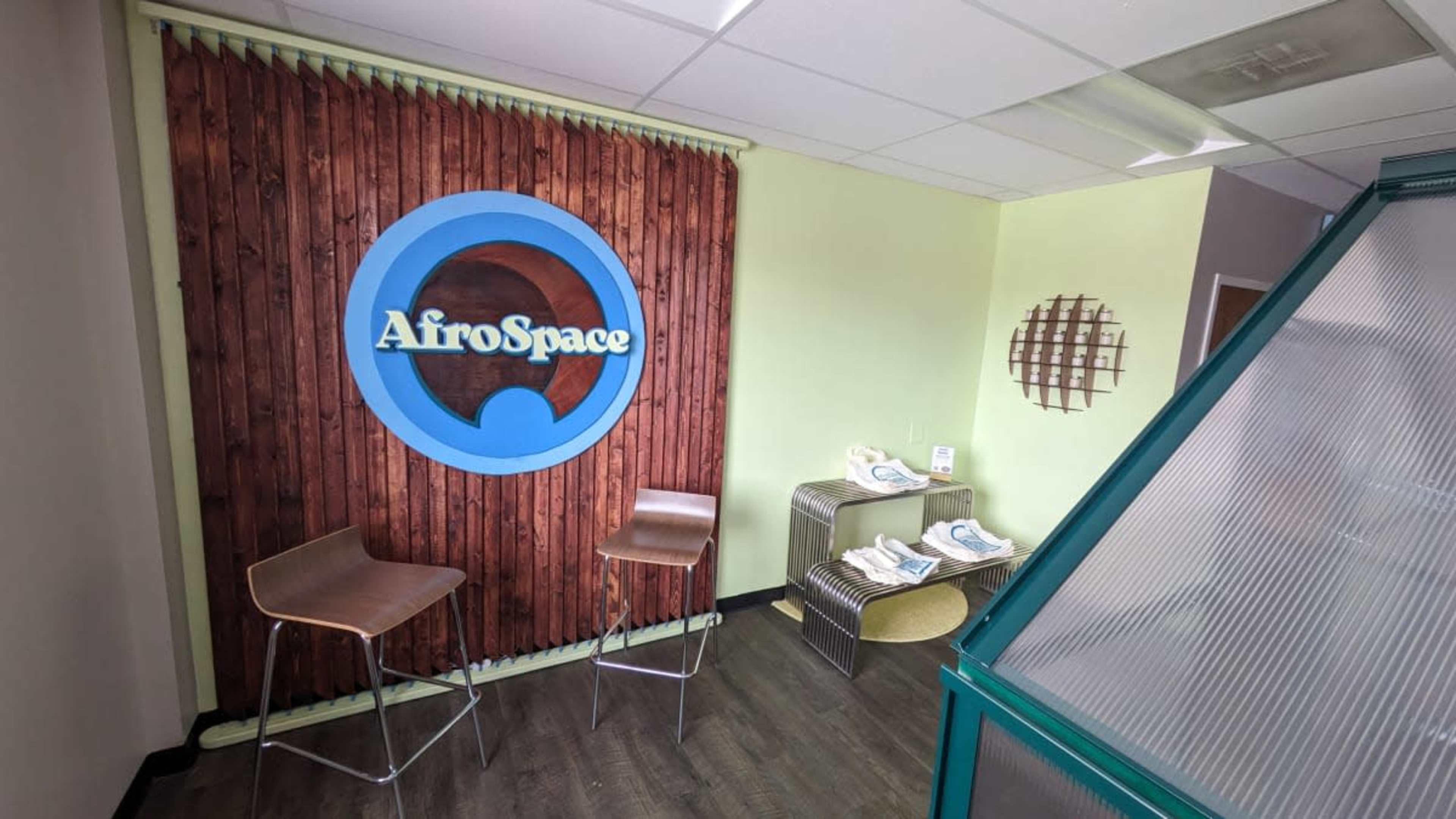 A modern interior space with a wooden accent wall featuring the logo "AfroSpace" and two bar stools in front of it, along with a display table on the side.
