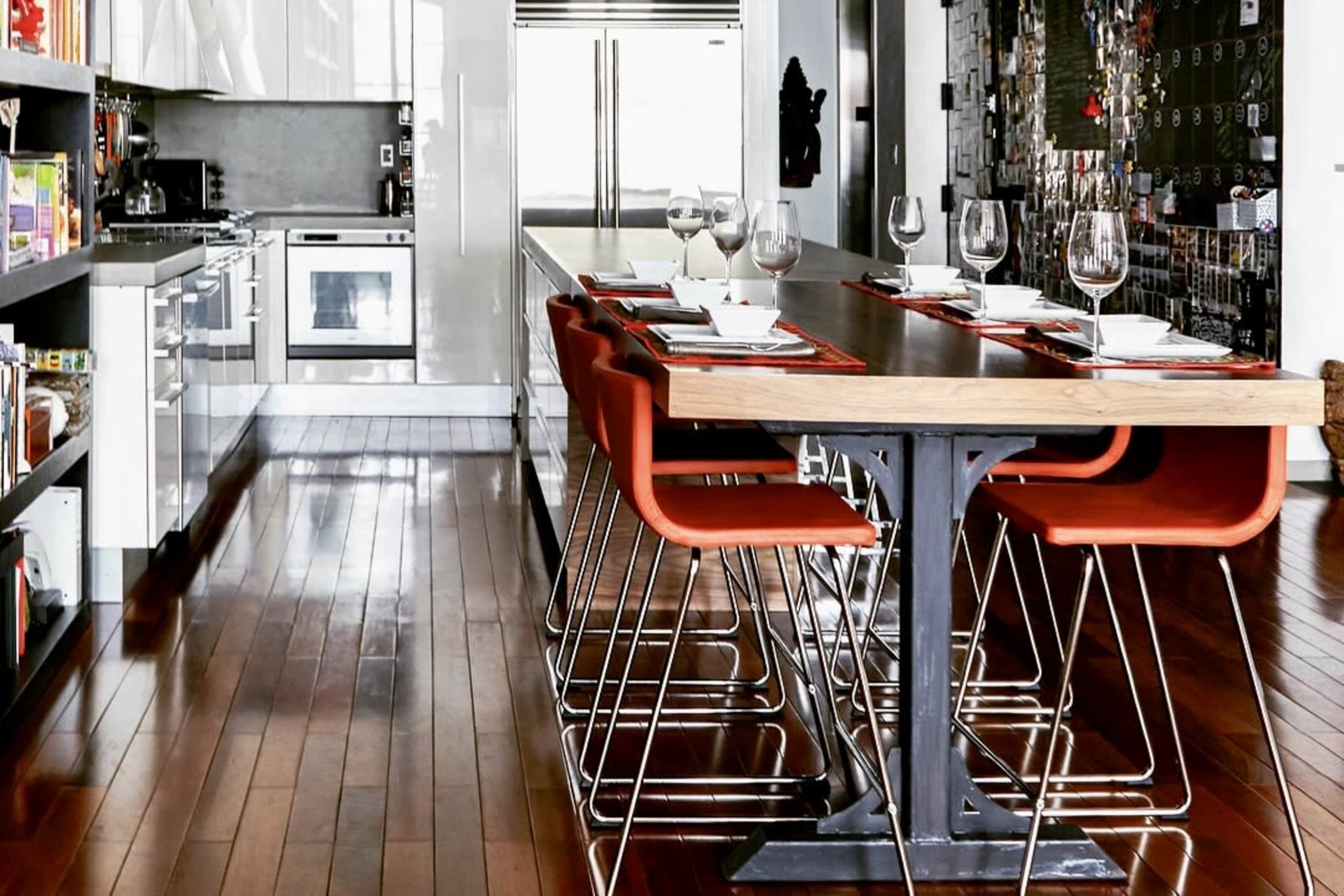 A modern kitchen features a long wooden dining table with red chairs and a well-organized cooking area in the background.